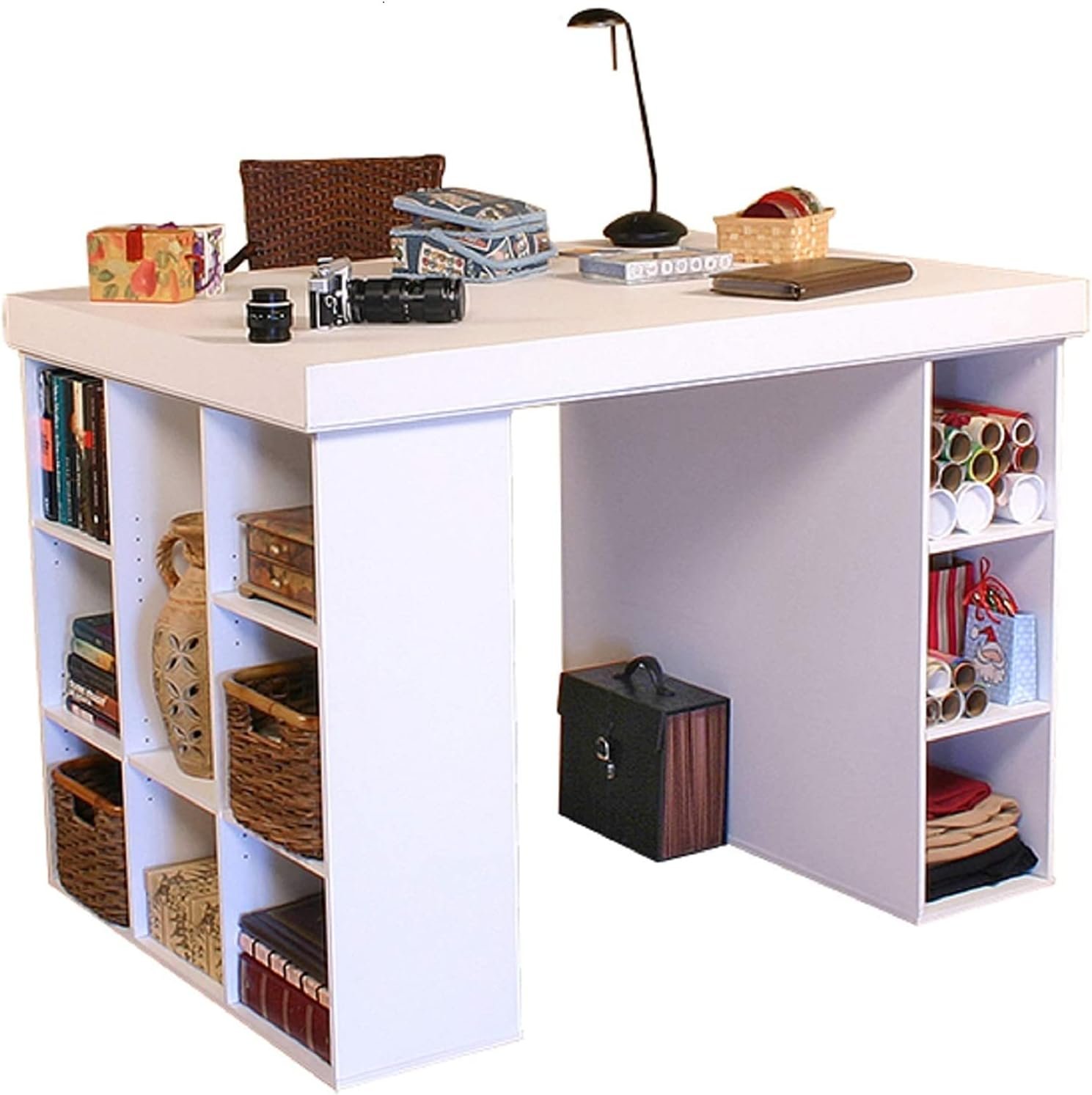 A white L-shaped desk with various items on its surface, including a table lamp, notebooks, a camera, and boxes. The desk has open shelving on both sides filled with books, baskets, and other supplies.