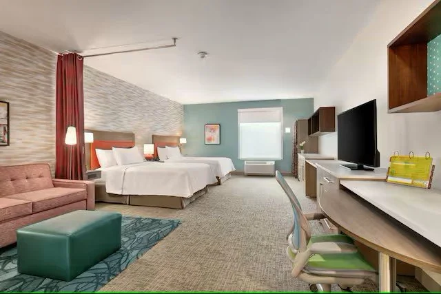 Hotel room with two beds, a pink sofa, a green ottoman, a desk with a chair, a TV, and artwork on the wall.