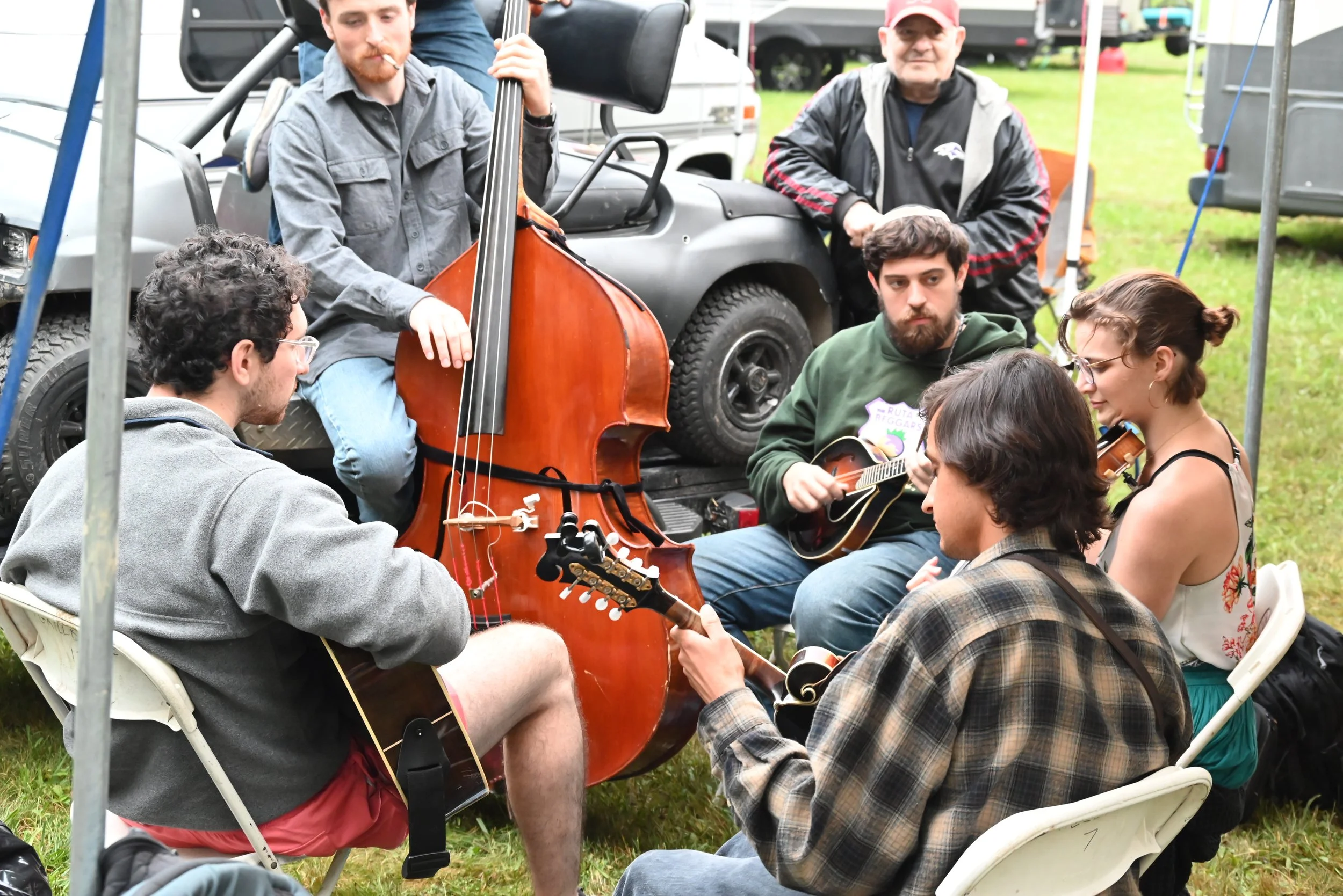 A group of six people playing musical instruments outdoors, with some seated and others standing, near a vehicle and tents on a grassy area.