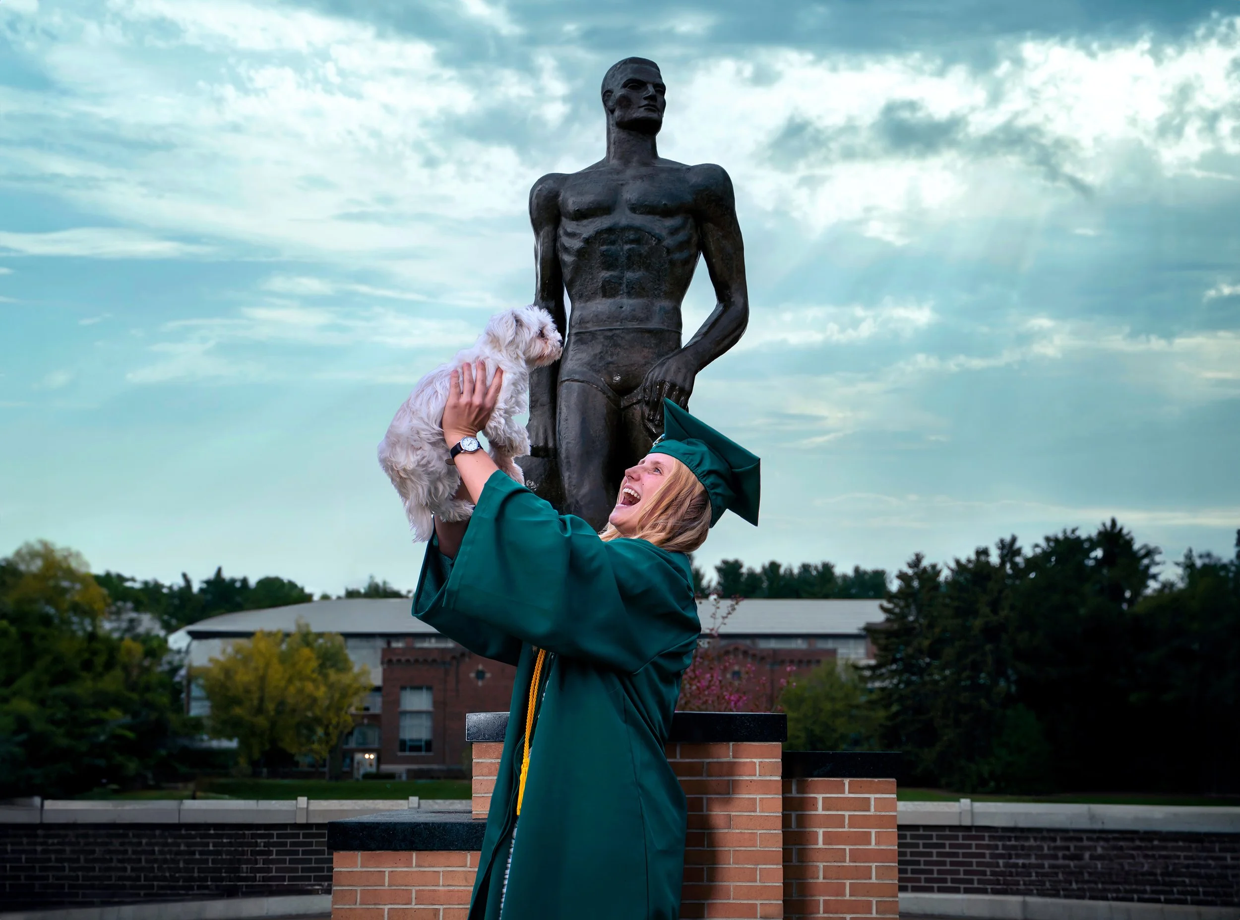 A joyful MSU graduate in her cap and gown sharing a special moment with her dog on campus in East Lansing, Michigan by the Spartan statue - celebrating years of hard work and a new beginning. Photography by AA Studios