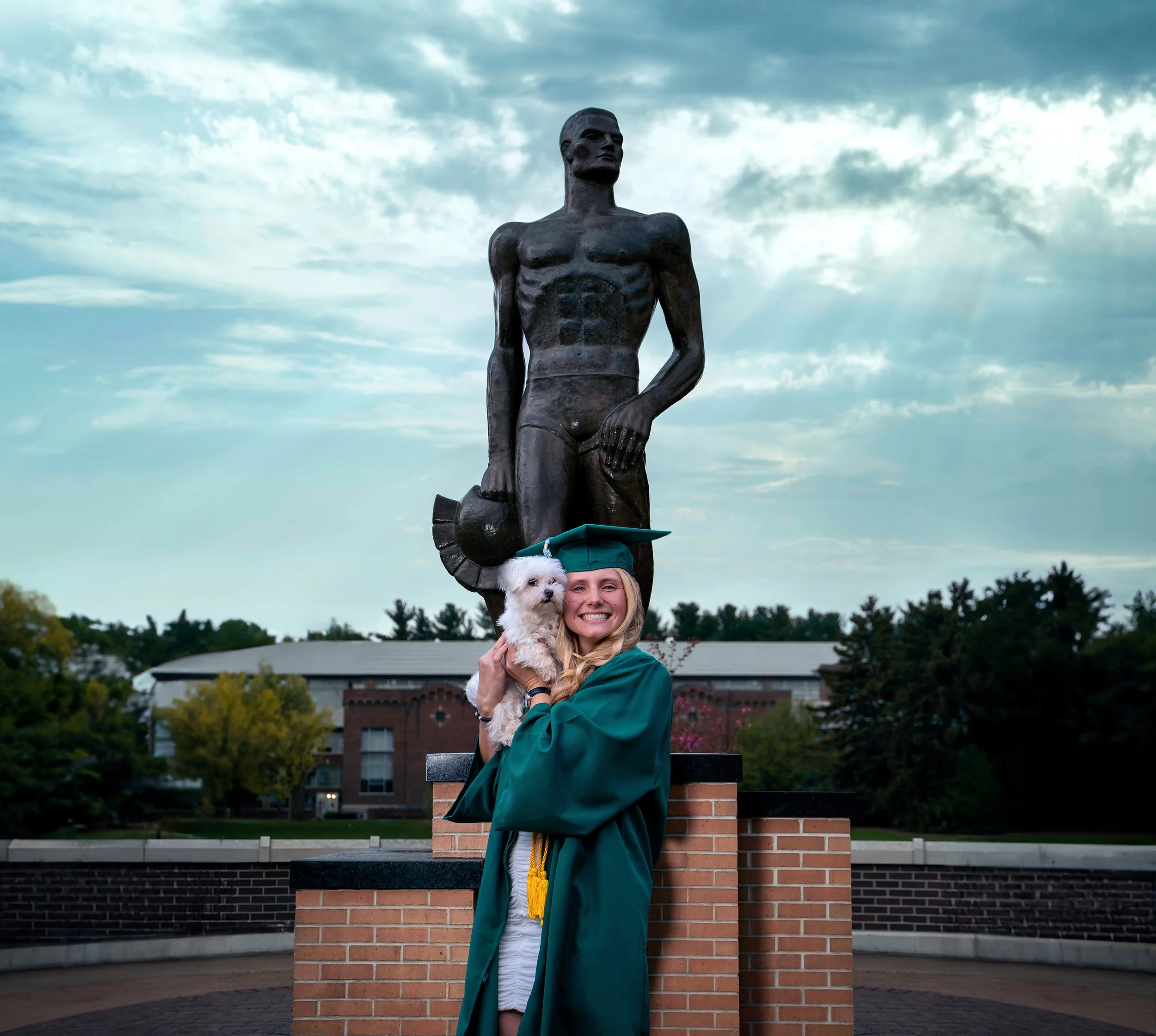 Michigan State University graduate celebrating with her dog during a cap and gown graduation session in East Lansing, Michigan. Captured by a Lansing-based senior graduation photographer - AA Studios.