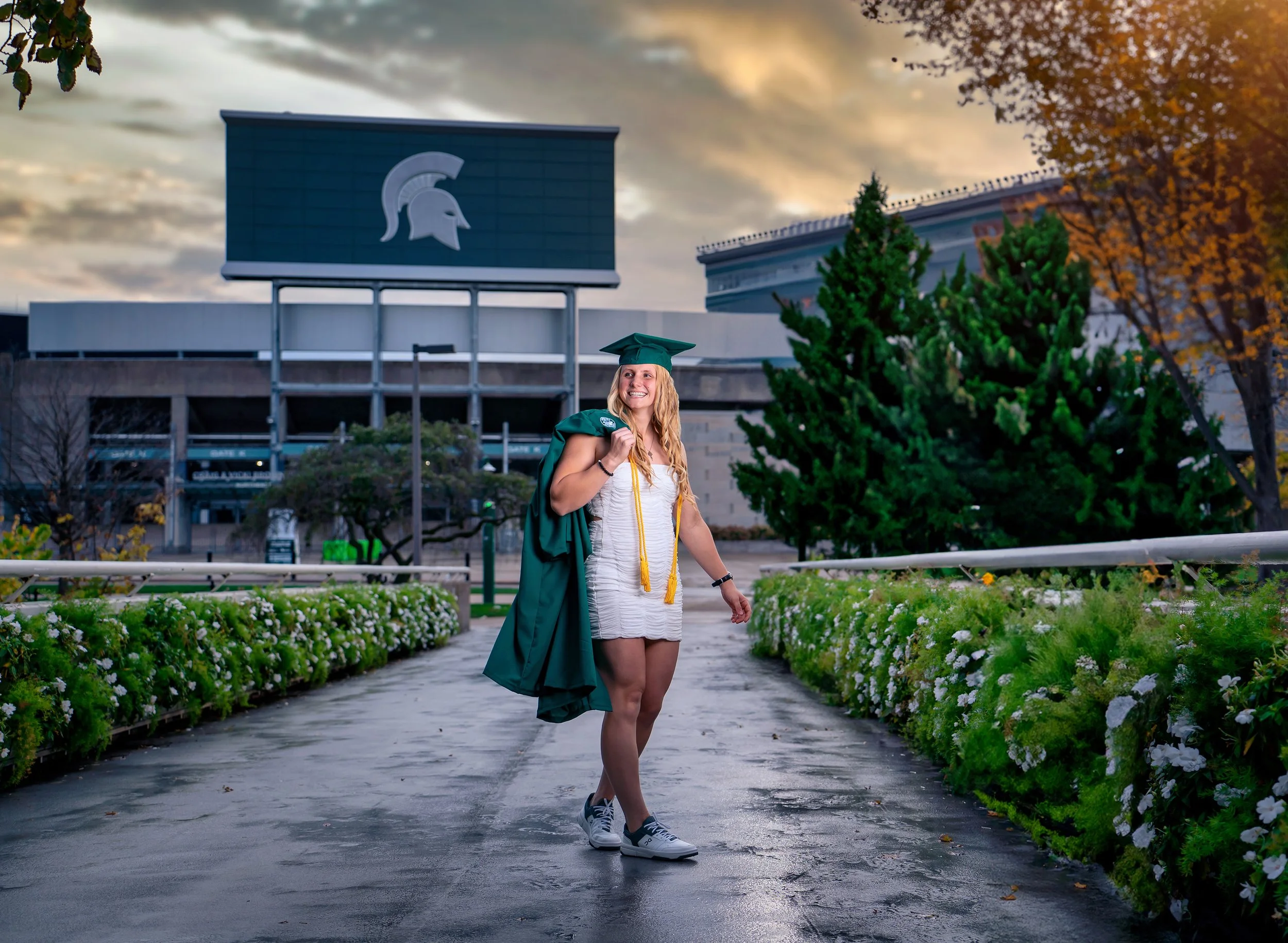 Michigan State University graduate walking across a campus bridge in cap and gown with the MSU football stadium sign in the background, captured in East Lansing, Michigan by AA Studios LLC