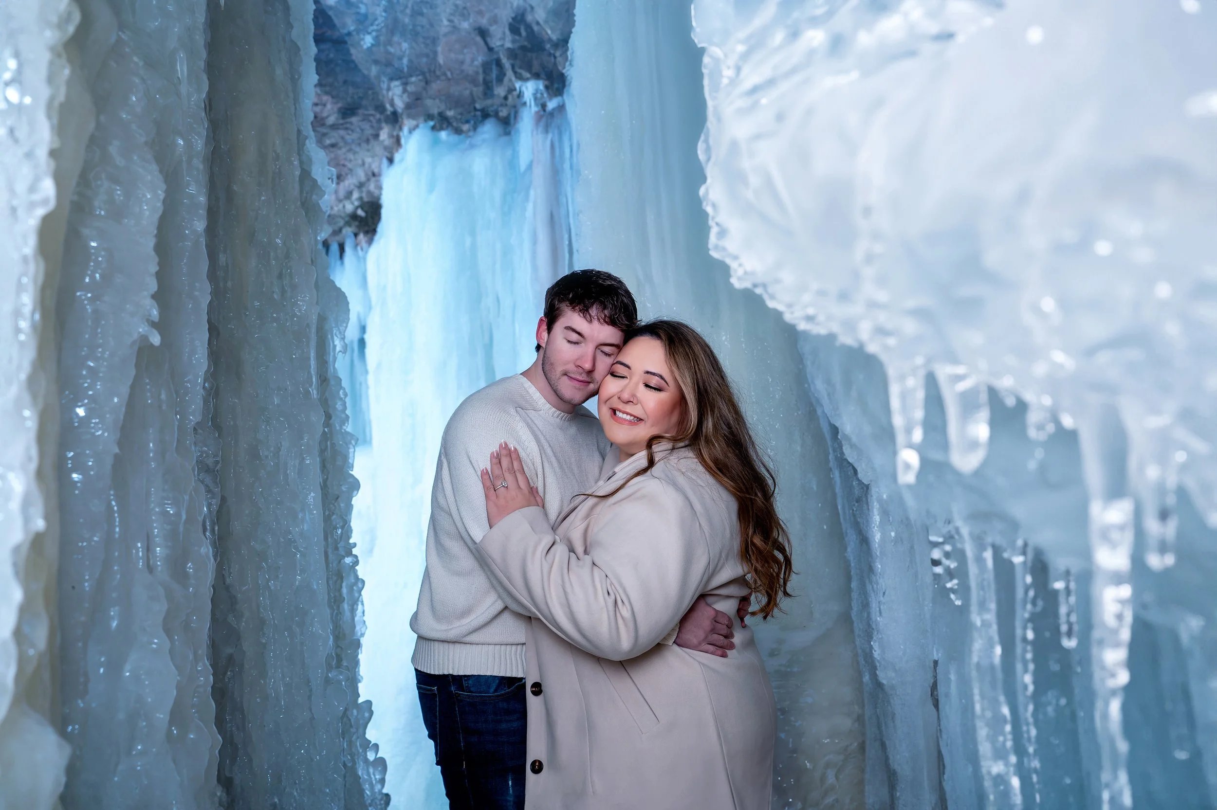 An ethereal winter engagement session unfolds at the frozen Wequiock Falls in Green Bay, Wisconsin, where a couple stands embraced behind the icy waterfall, surrounded by soft, dreamy light and a breathtaking winter landscape. Captured by AA Studios,