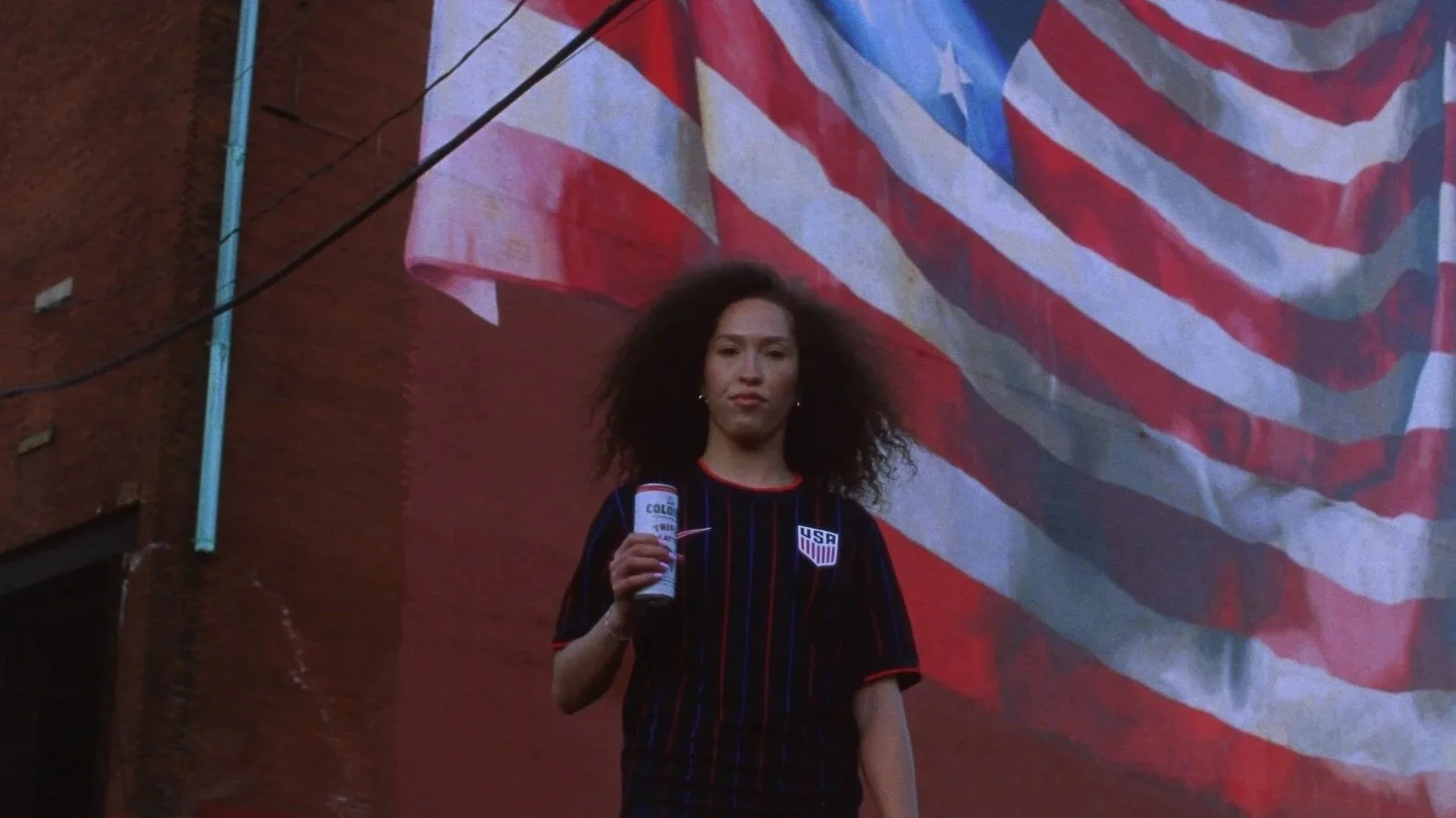 A stands confidently holding a can of La Colombe coffee, wearing a USA World Cup jersey. Behind them, a large American flag mural fills the background.