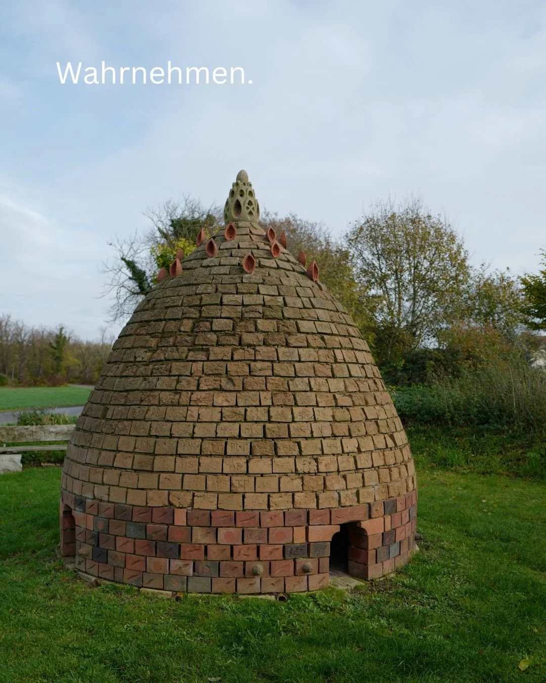 Ein rundes Haus aus Ziegelsteinen mit einer konischen Form, das wie eine große Höhle aussieht, im Freien auf einem grünen Rasen, mit Bäumen im Hintergrund, und der deutschen Schriftzug "Wahrnehmen" oben links.