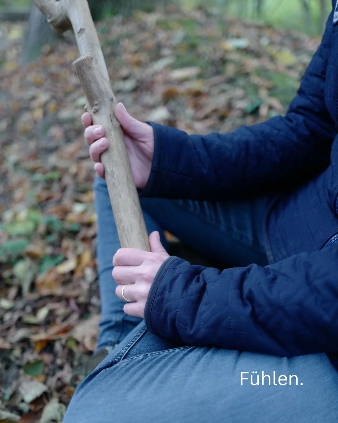 Eine Person sitzt im Wald, hält einen kleinen Ast in den Händen und fühlt ihn. Die Umgebung ist von Laub bedeckt, es ist herbstlich.