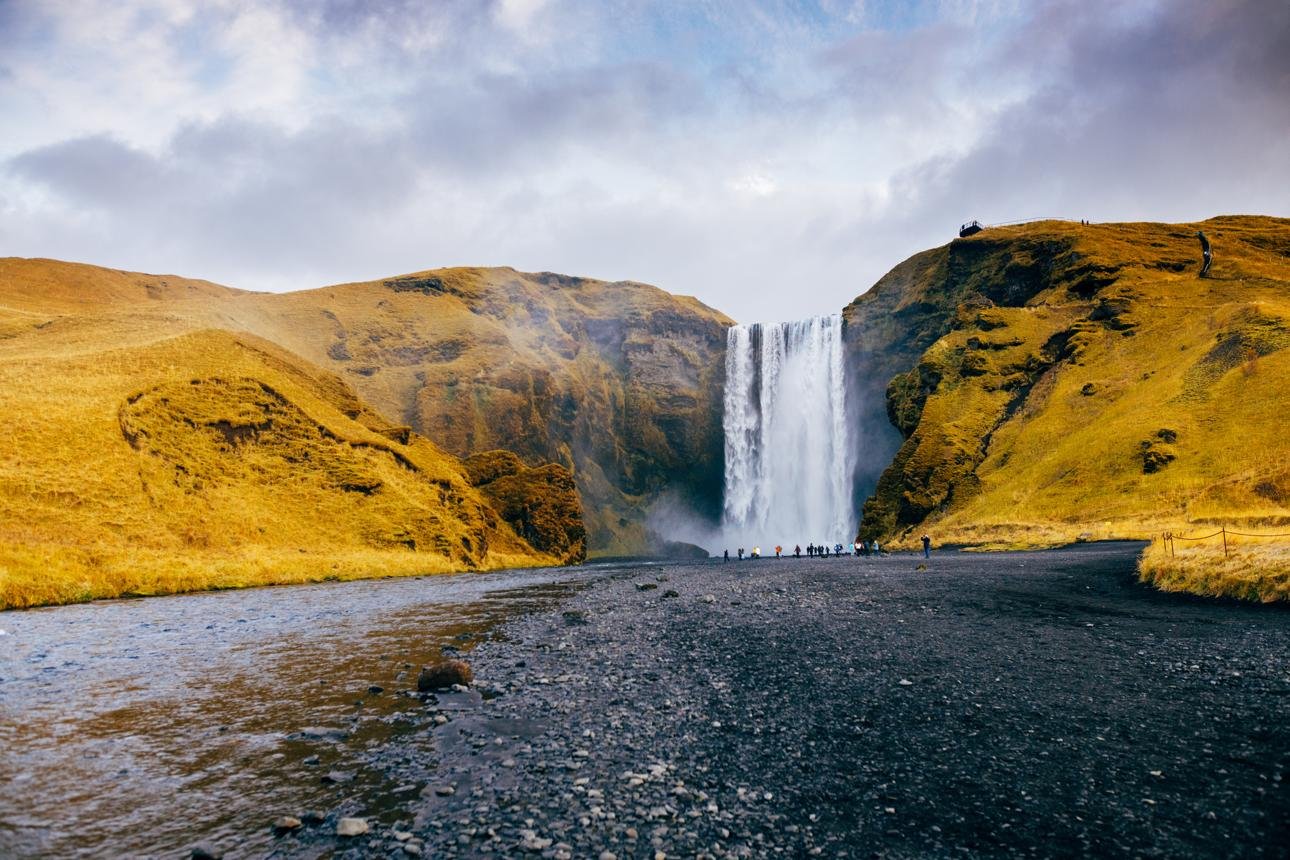 A large waterfall cascading into a black pebble beach with a river flowing in the foreground. Green grassy hills surround the waterfall, with a cloudy sky above. Small figures of tourists are visible near the base of the waterfall.