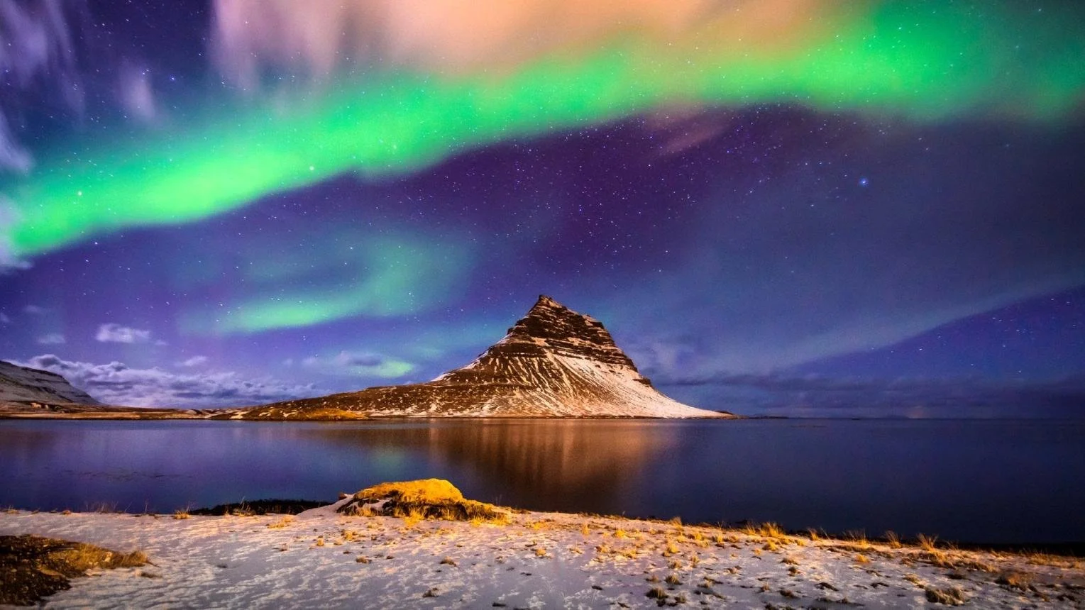 Nighttime scene of Kirkjufell mountain reflected on water, with colorful northern lights in the sky, some snow on ground. Iceland group trips for solo travelers.