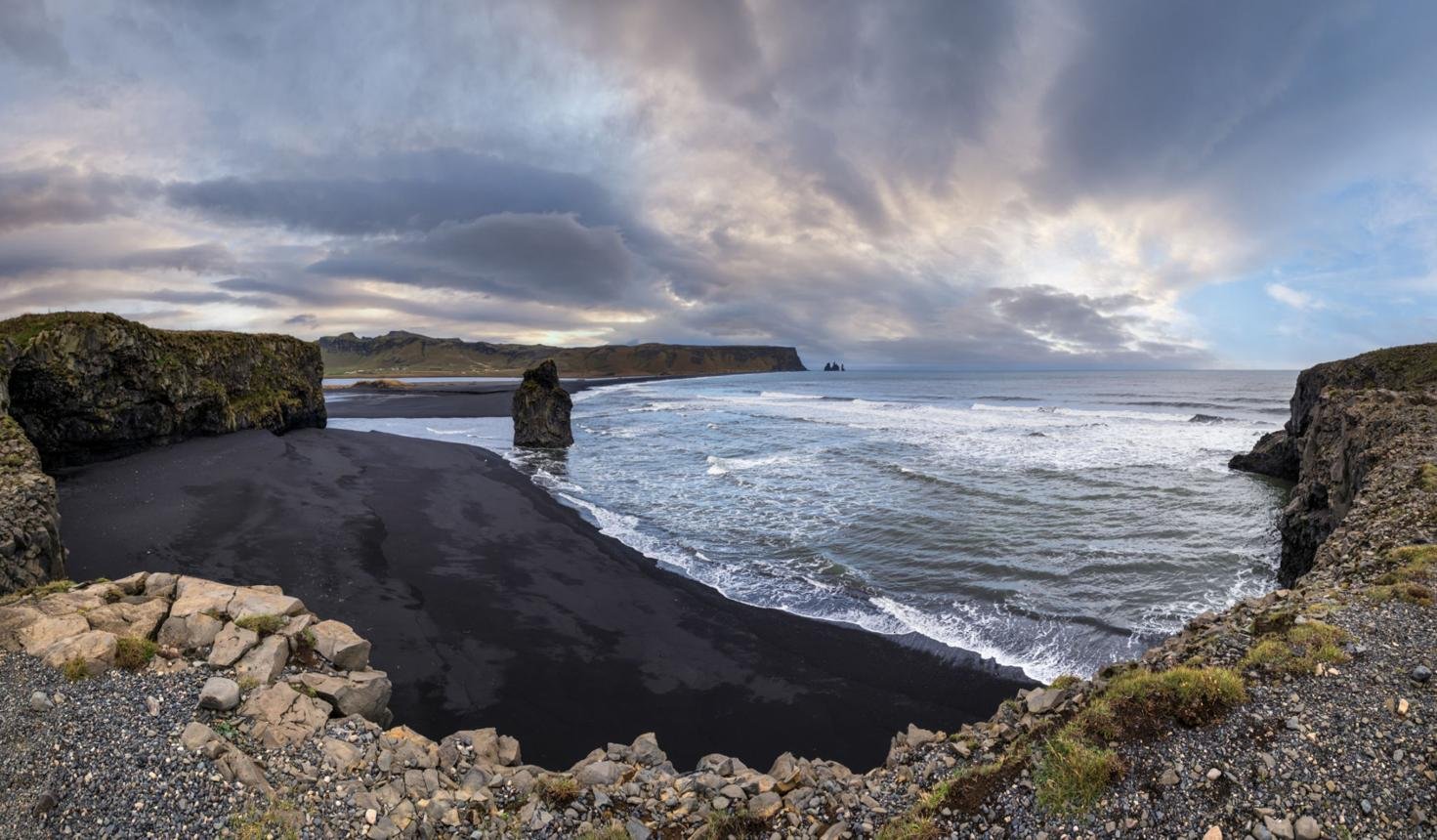 A rugged black sand beach with large rocks and dramatic cliffs under a cloudy sky.