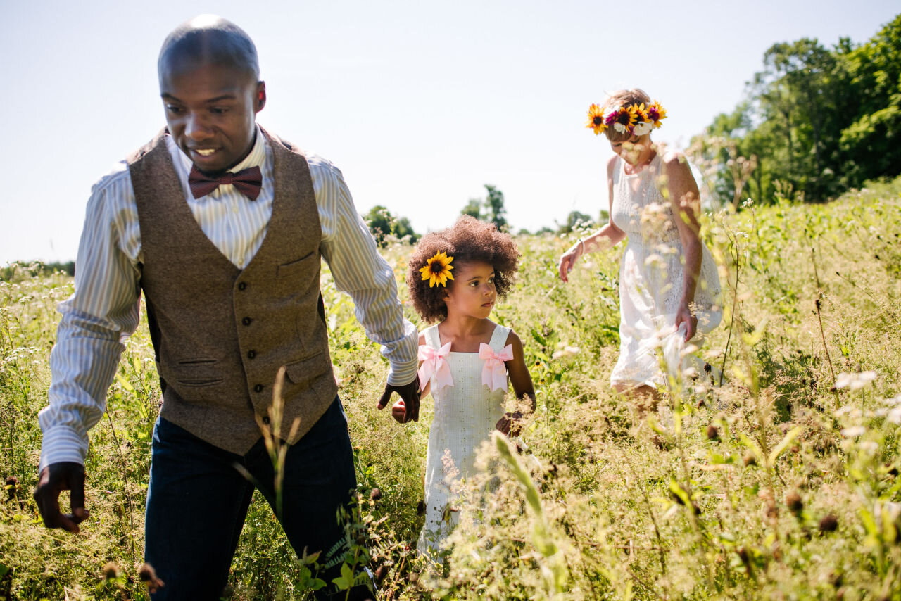 Groom, bride and child walking through a field of wild flowers at their elopement near Kingston, Ontario