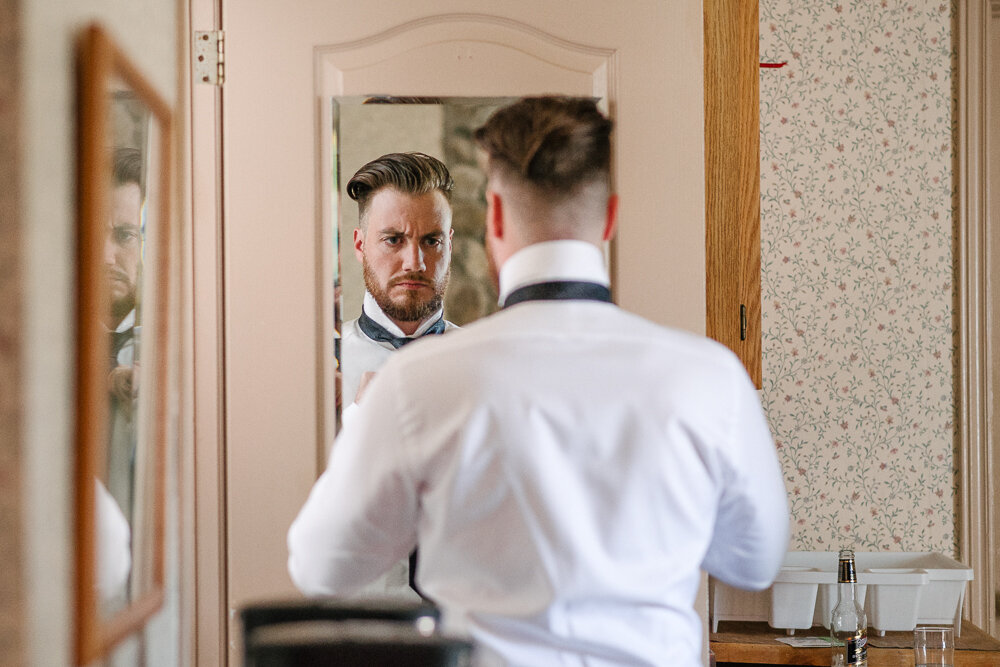candid photo of Groom tying his tie in the bathroom mirror