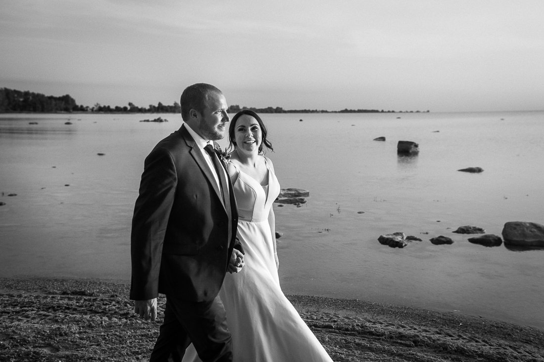 Black and white image of bride and groom walking hand in hand kissing near the beach. 