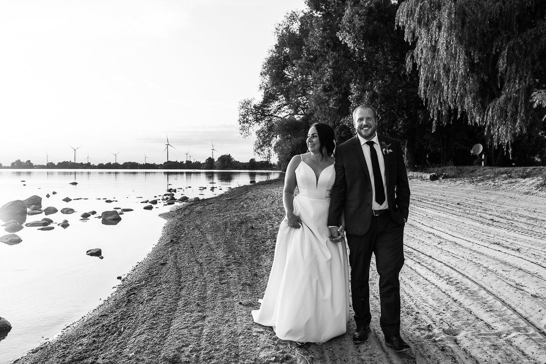 Black and white image of bride and groom walking hand in hand kissing near the beach. 