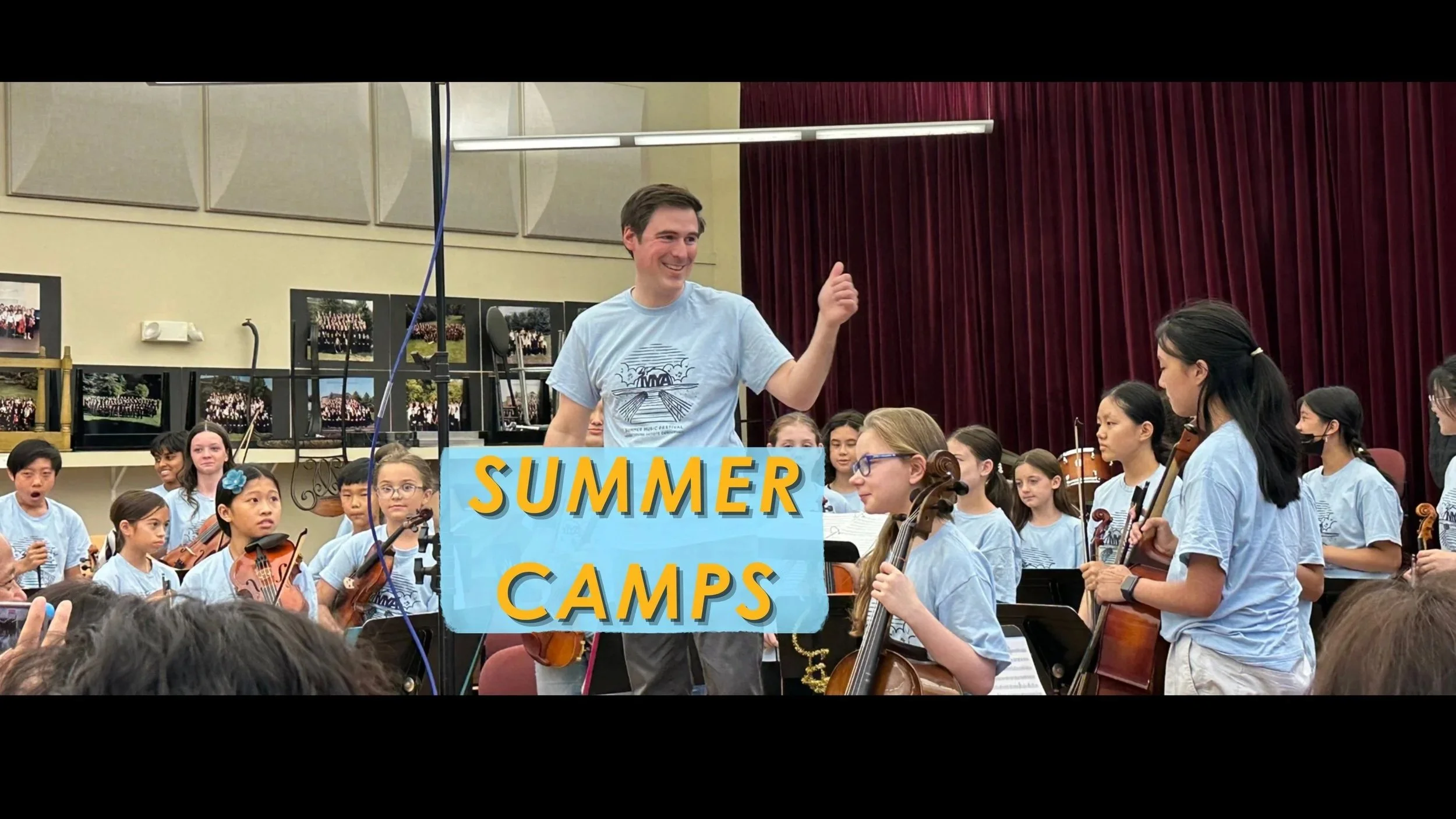 A conductor smiles and waves his hand at an orchestra full of young musicians, all of whom are wearing blue matching shirts and smiling