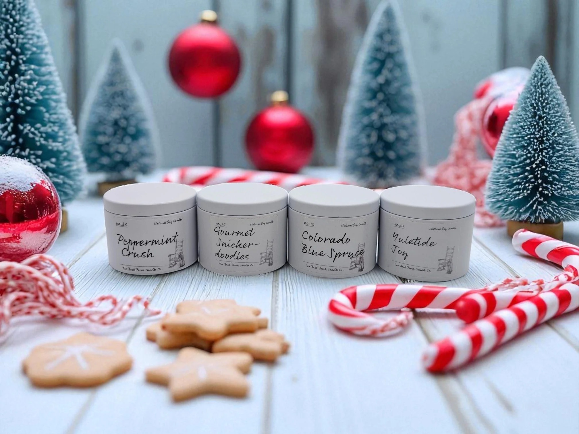 Four white candles with handwritten labels, surrounded by Christmas decorations including candy canes, snow-dusted miniature trees, red ornaments, cookies, and red and white striped string on a white wooden surface.