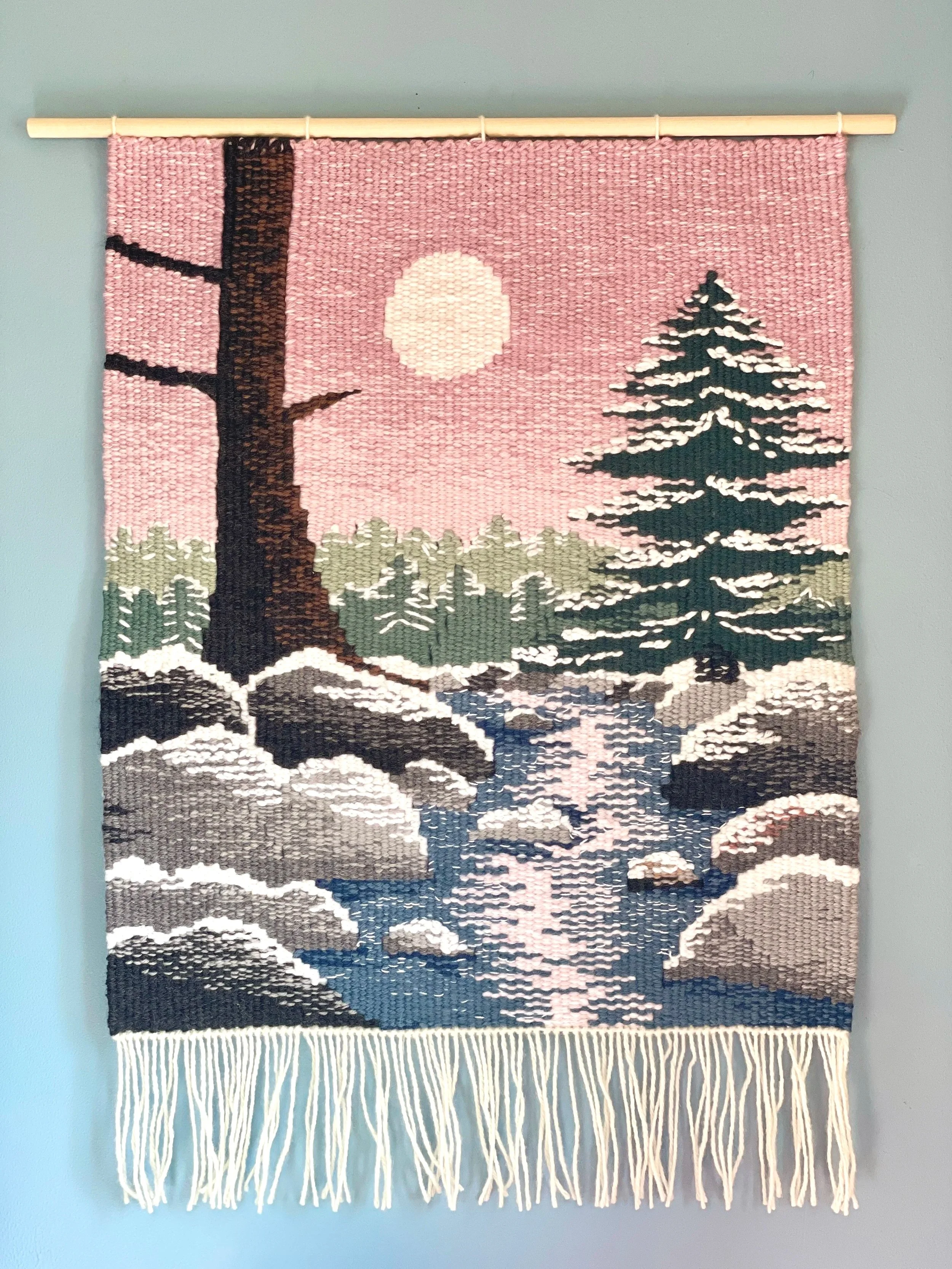 A tapestry showing a winter scene with trees, a river, and a full moon, by Emily Wick.