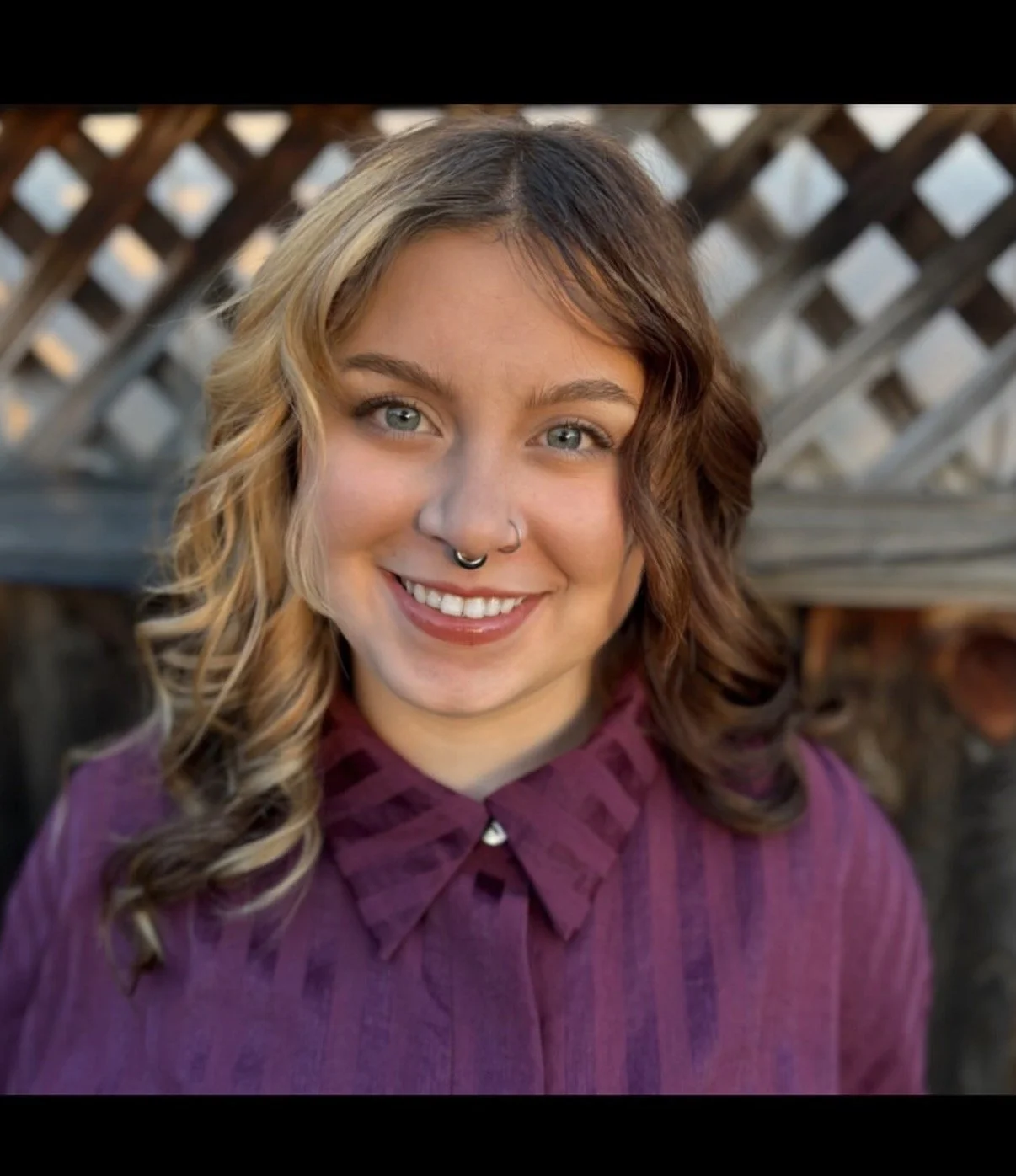 A young woman with curly hair, light skin, and blue eyes, smiling, wearing a purple shirt with a ruffled collar and a nose piercing, outdoors with a wooden fence in the background.