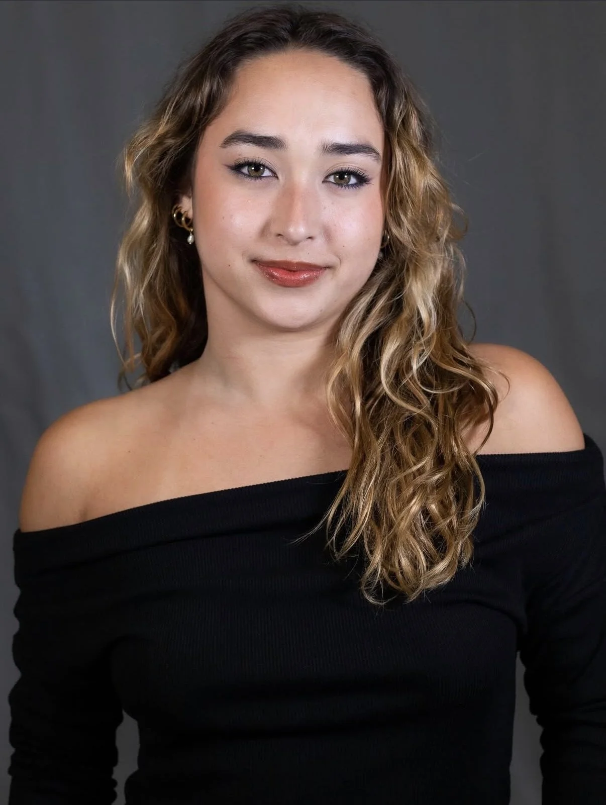Portrait of a young woman with wavy brown hair wearing a black off-the-shoulder top and earrings, smiling against a gray background.