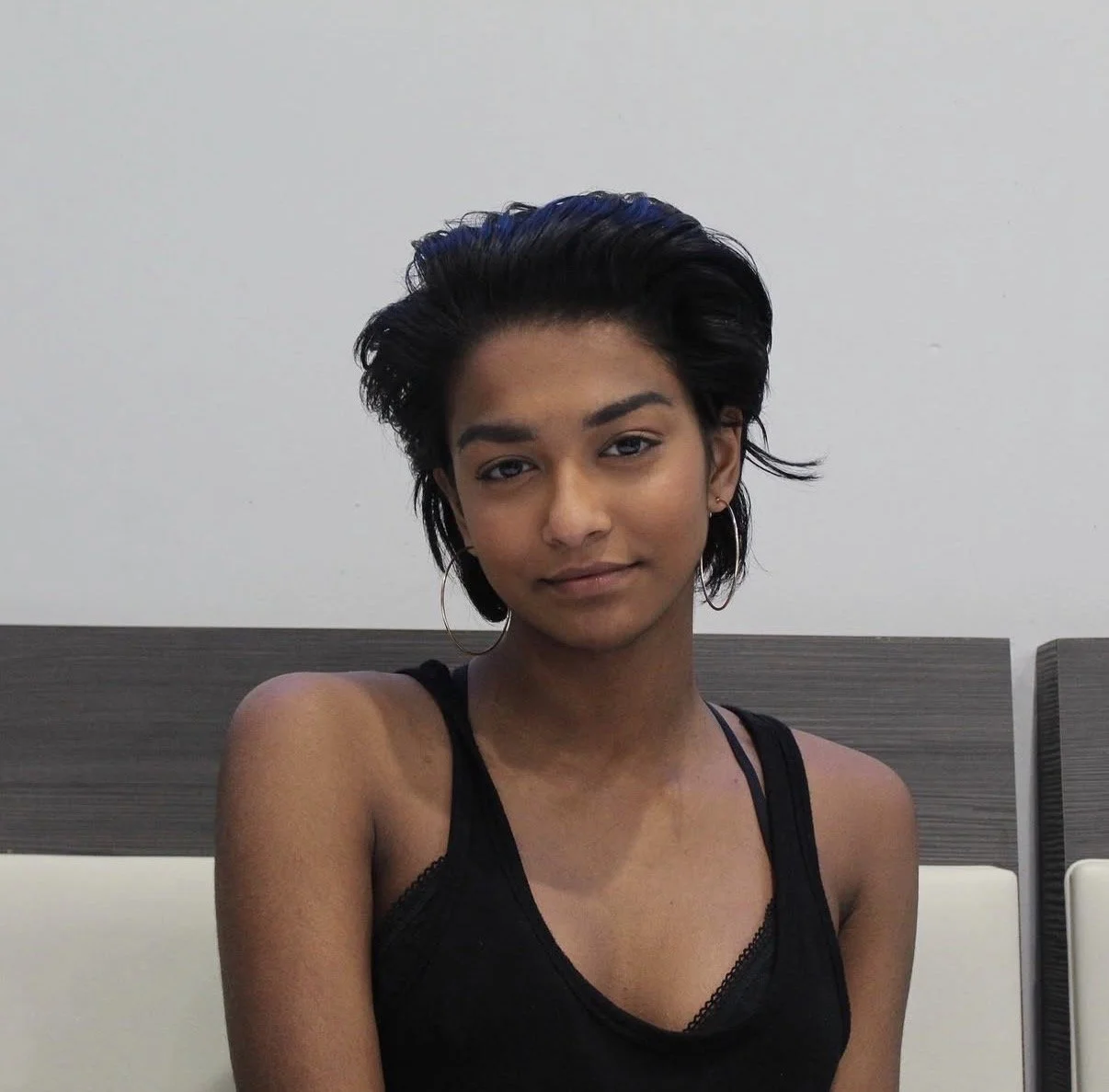 A young woman with short, dark hair, wearing hoop earrings and a black sleeveless top, sitting in front of a plain wall and a dark wooden headboard.