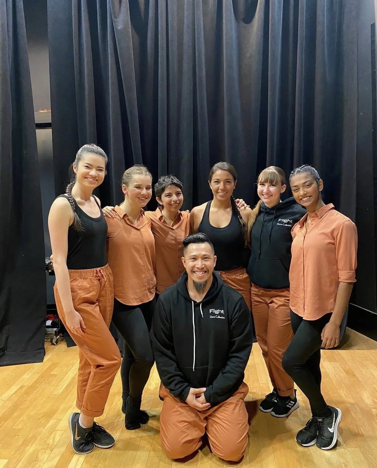 Group of seven dancers, six women and one man, smiling and posing in a dance studio with black curtains and wooden floor. They are wearing dance attire with some in black and others in orange-brown pants and tops, and the man is kneeling in front wearing a black hoodie with 'Flight' written on it.