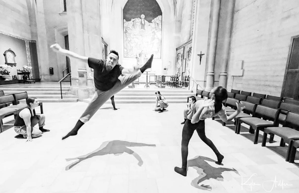 A person is performing a mid-air dance leap in a church or cathedral while others are seated or crouching on the floor around them, with a large religious painting and a cross on the wall in the background.