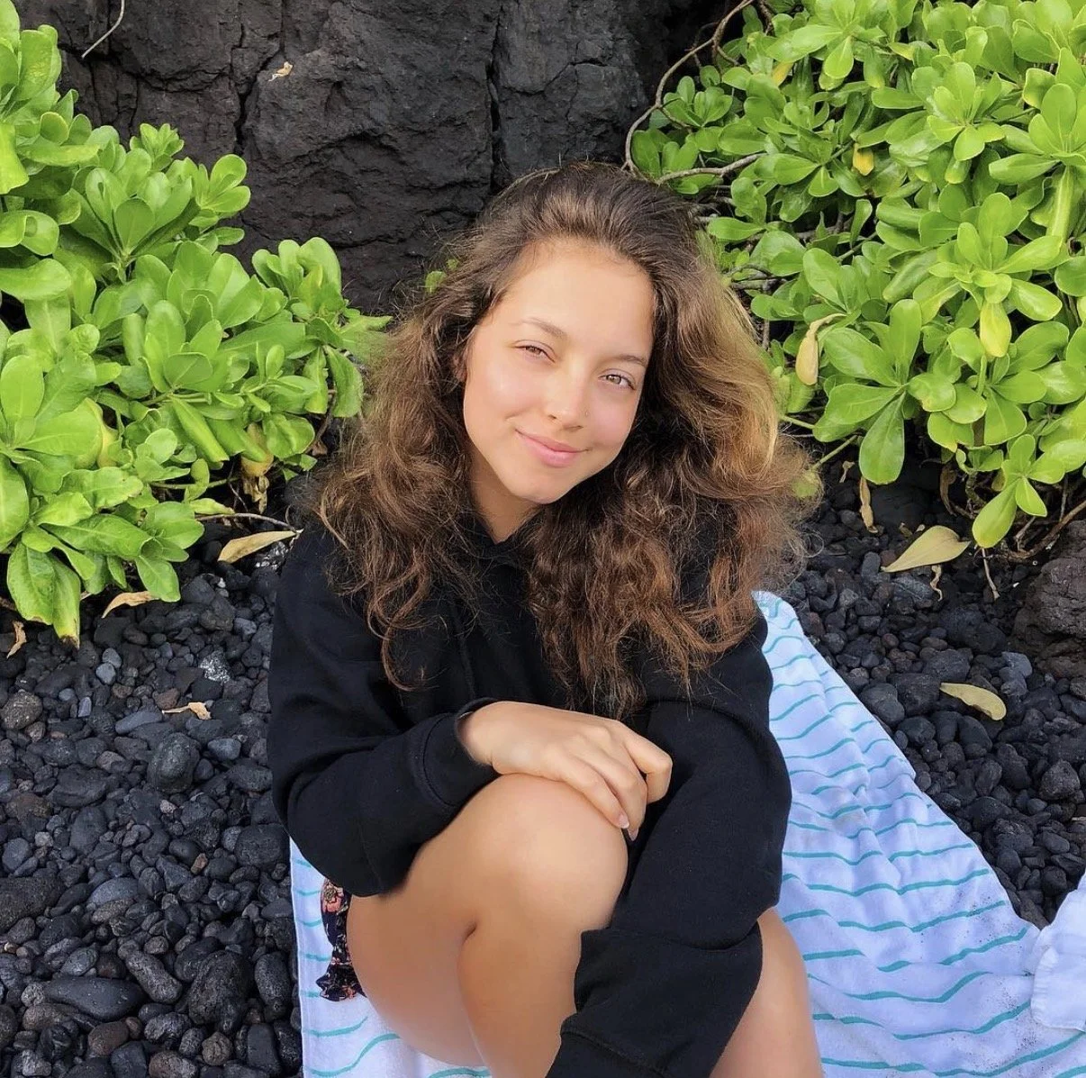 Young woman with wavy brown hair sitting on a striped towel on black volcanic rocks, surrounded by green leafy plants, smiling softly.