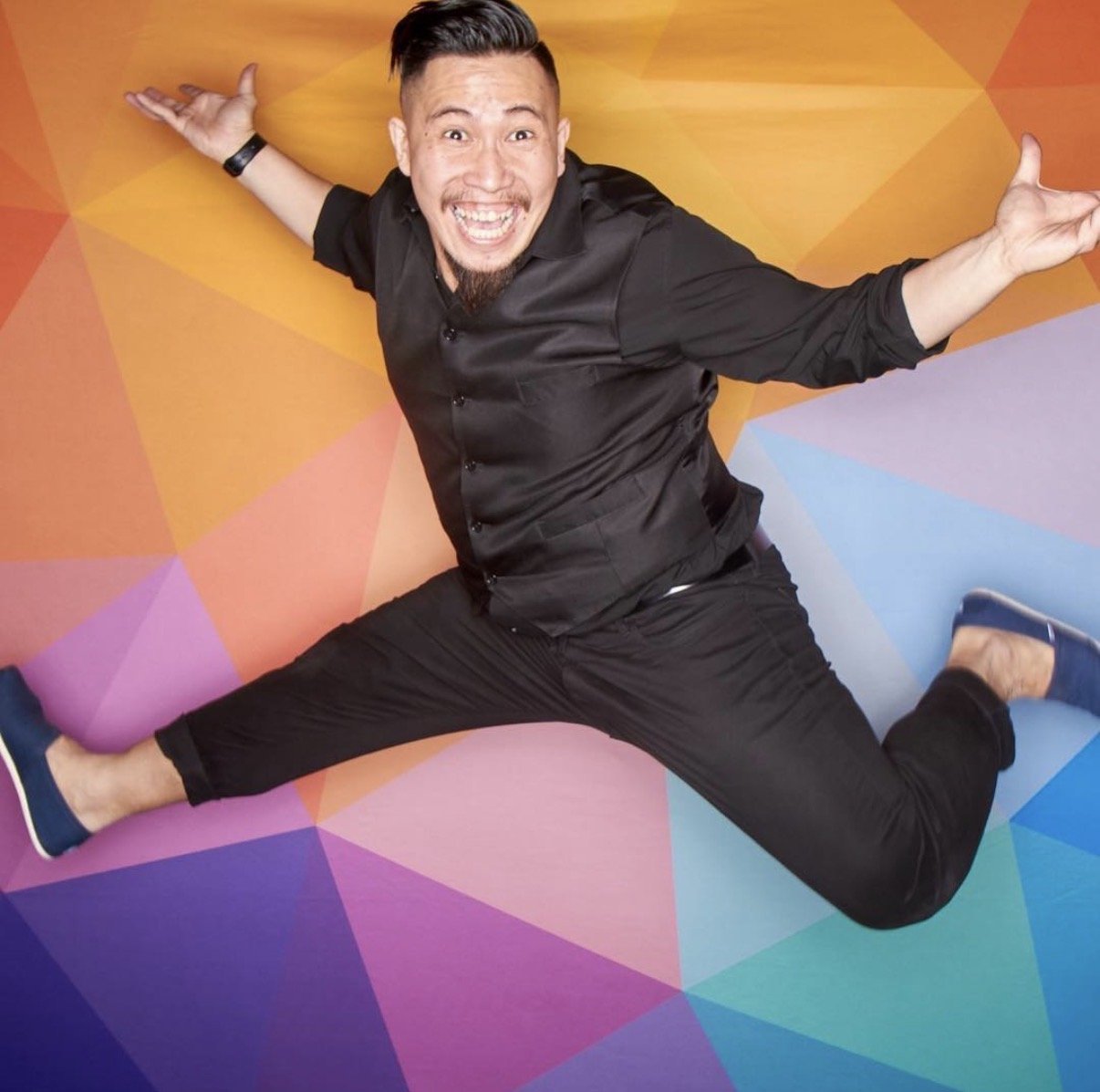 A man smiling widely with arms outstretched, doing a split on a colorful geometric background.
