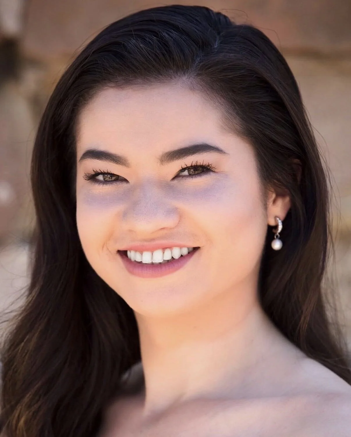 Close-up portrait of a smiling woman with long dark hair, wearing pearl earrings, outdoors in natural light.