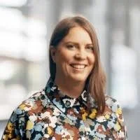 Hän hymyilee ja katsoo kohti kameraa, taustalla ikkunat ja rakennukset