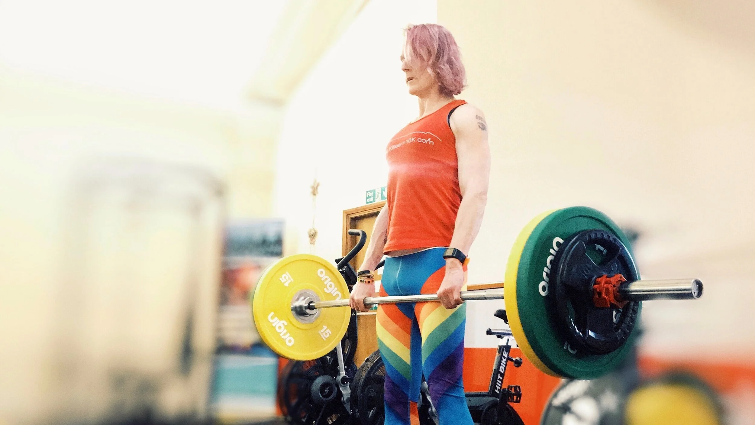 A person in rainbow leggings and a red vest stands holding a barbell with olympic plates on it.