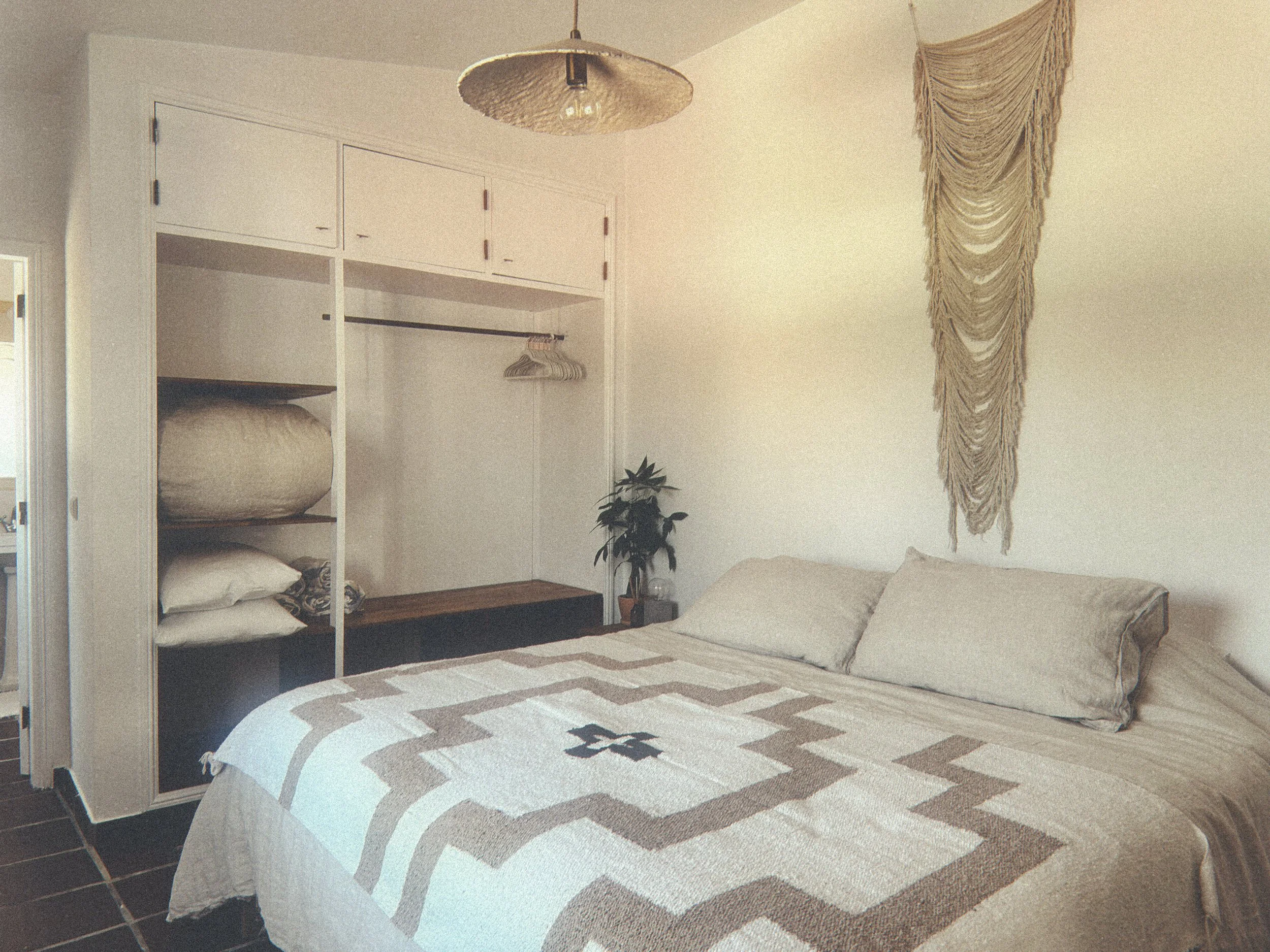 A cozy bedroom featuring a white bed with a beige and black geometric patterned quilt, a wooden nightstand with a potted plant, and an open closet with shelves and a hanging rod. A hanging light fixture is above the bed, and a decorative beige macrame wall hanging is on the wall.