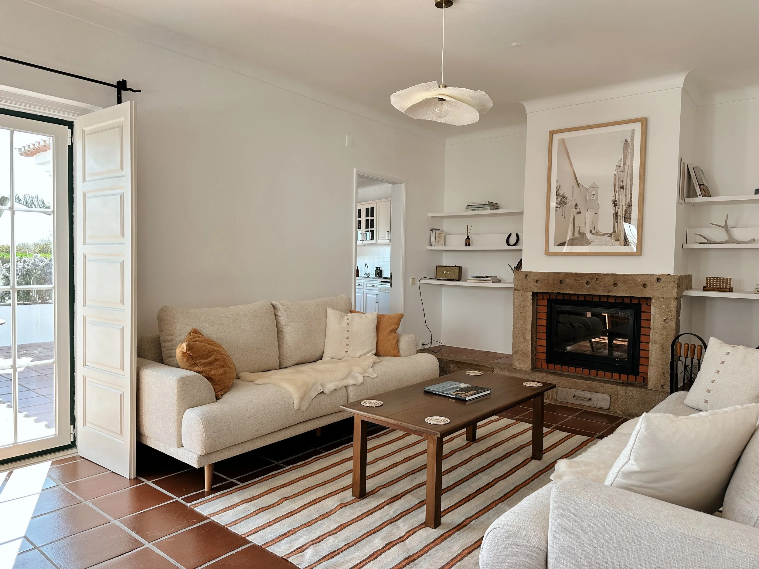 Living room with a fireplace, white walls, built-in white shelves with books and decorative objects, a beige sofa with a pillow, a wooden coffee table with a plant, and an ottoman near the fireplace.