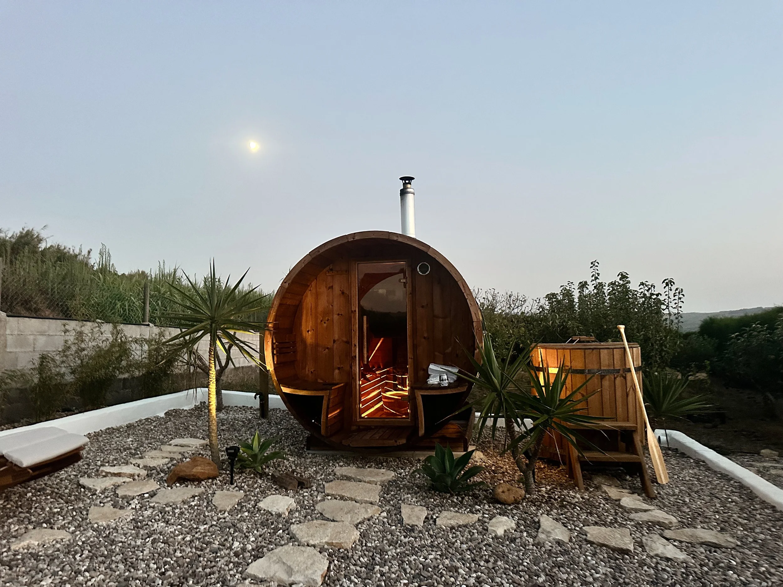 A wooden barrel-shaped outdoor sauna with a small staircase leading to the entrance, surrounded by desert plants and a gravel pathway, set in a natural landscape under a pale sky with the moon visible.