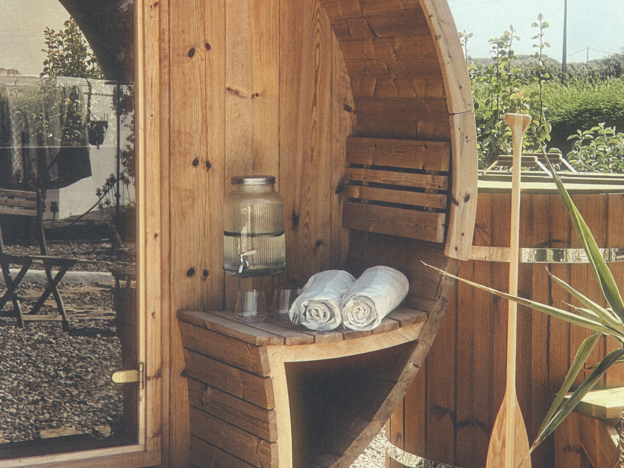 Wooden outdoor shower with a curved design, featuring a glass door, a water barrel with a tap, folded towels, and small glasses on a wooden bench.