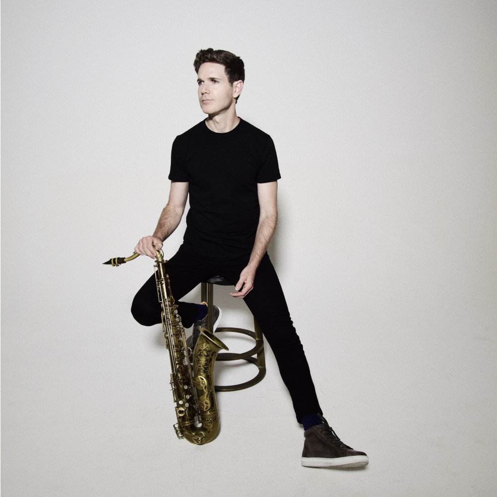 Un uomo con maglietta nera e jeans neri seduto su uno sgabello, tiene un sassofono d'oro, si trova di fronte a uno sfondo bianco.