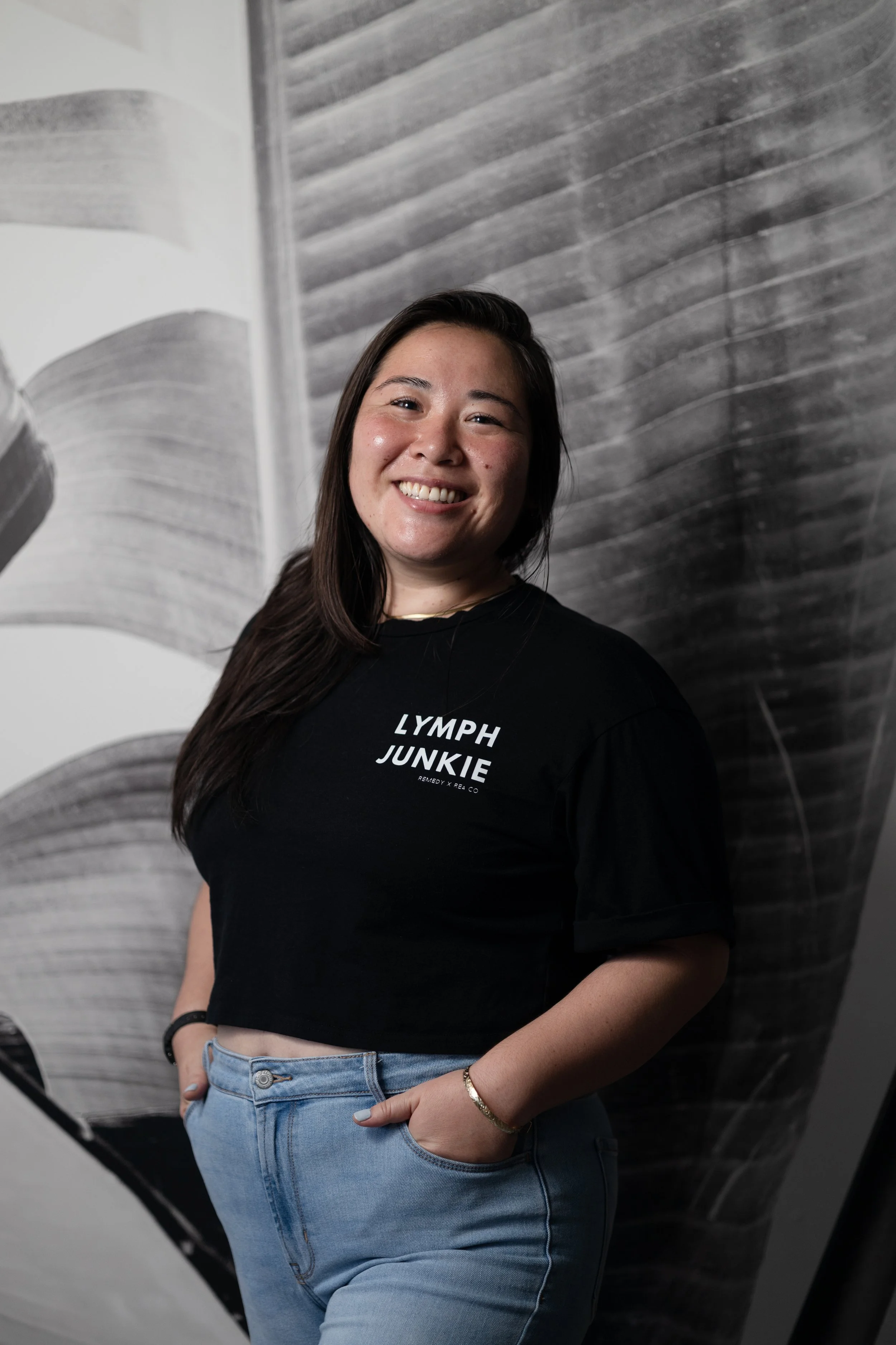 A woman smiling with long dark hair, wearing a black crop top with 'LYMPH JUNKIE' written on it, and light blue jeans, standing in front of a black and white abstract background.