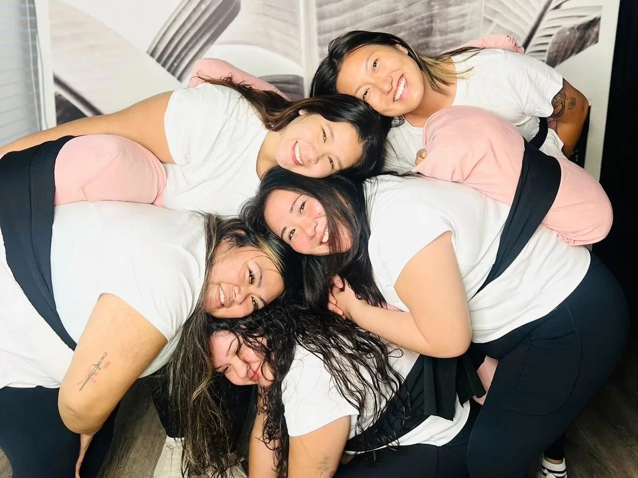 Five women lying on a bed, smiling and hugging each other.