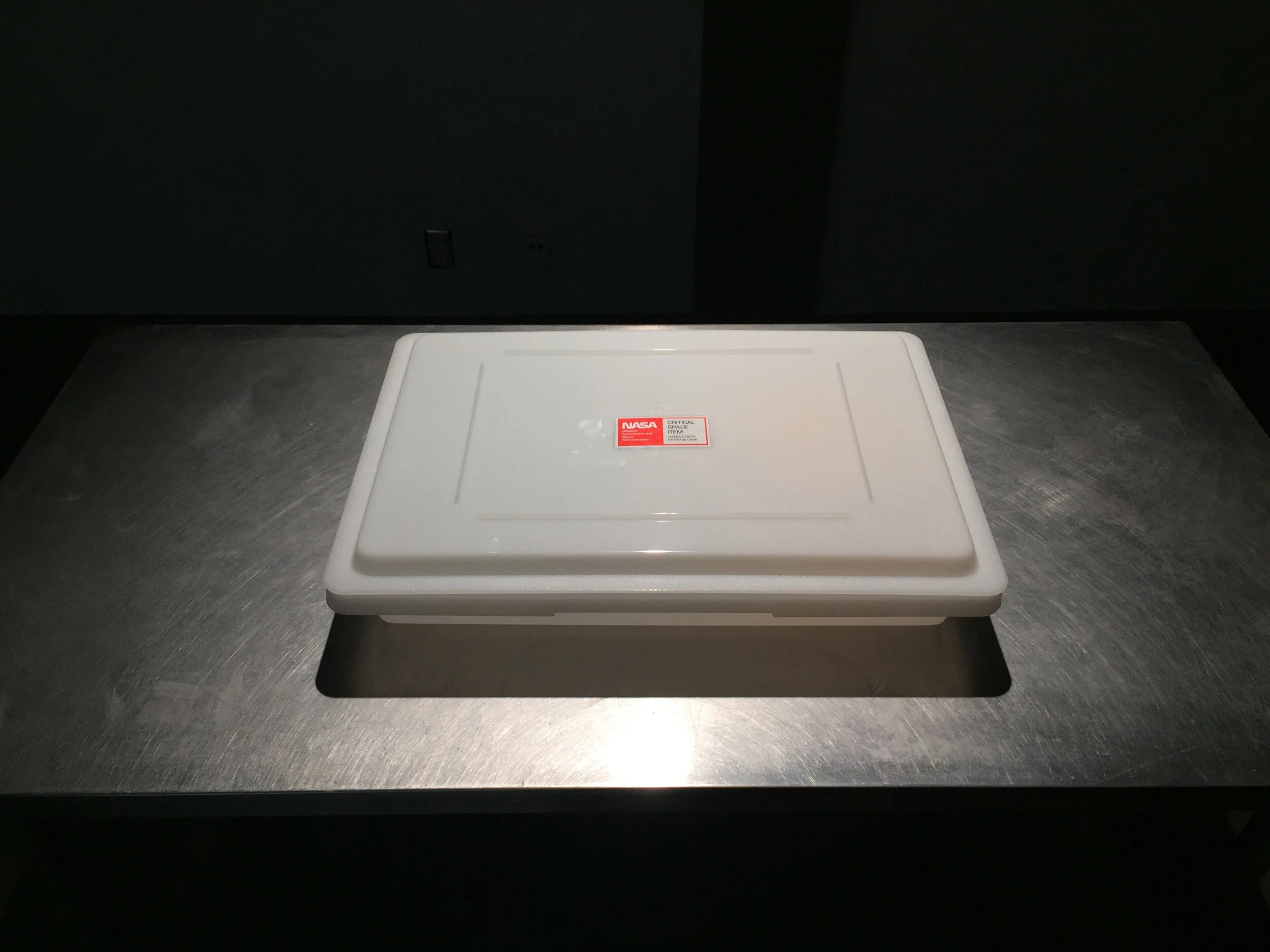 Michael Edward Miller Critical Space Item , 2018, dyed slip cast fired clay powder, laser cut coloured plexiglass, plastic catering container, authentic NASA critical space item label, altered authentic NASA non-disclosure agreements, respiratory masks, hypoallergenic gloves, stainless steel tables, plastic quarantine sheets, actors, participants