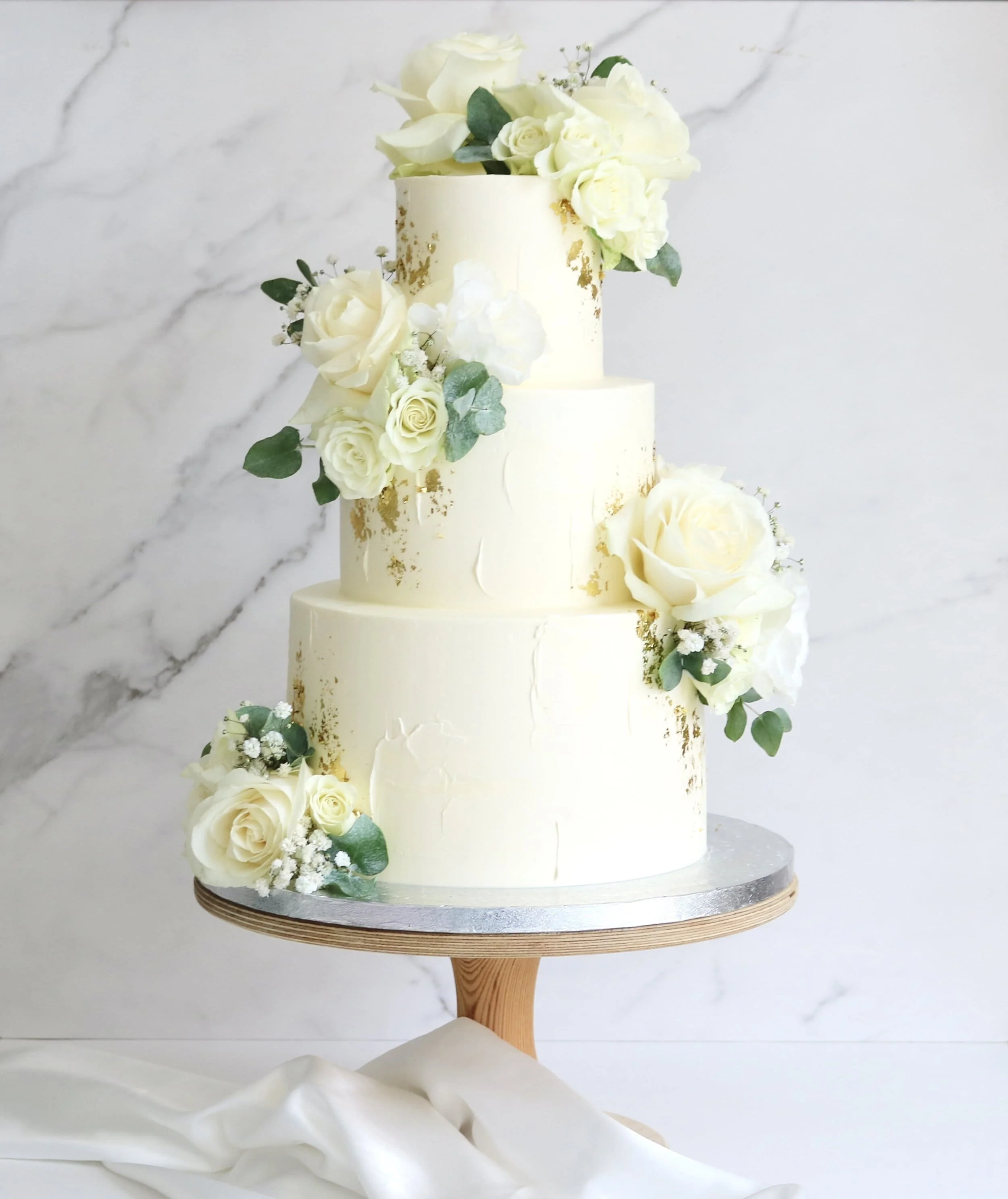 Three-tier white wedding cake decorated with pink and peach roses, pink carnations, and green eucalyptus leaves, set against a white marble background.