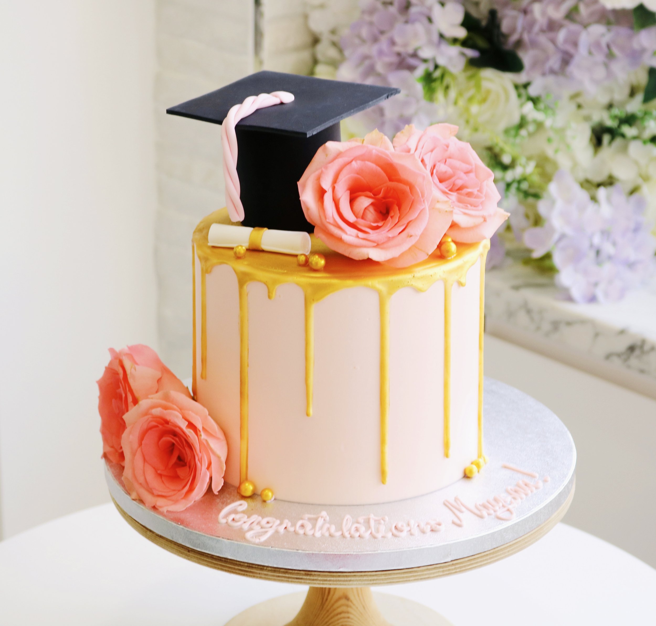 Graduation cake with edible hat and stroll, decorated with gold chocolate drips and fresh flowers and gold pearls.