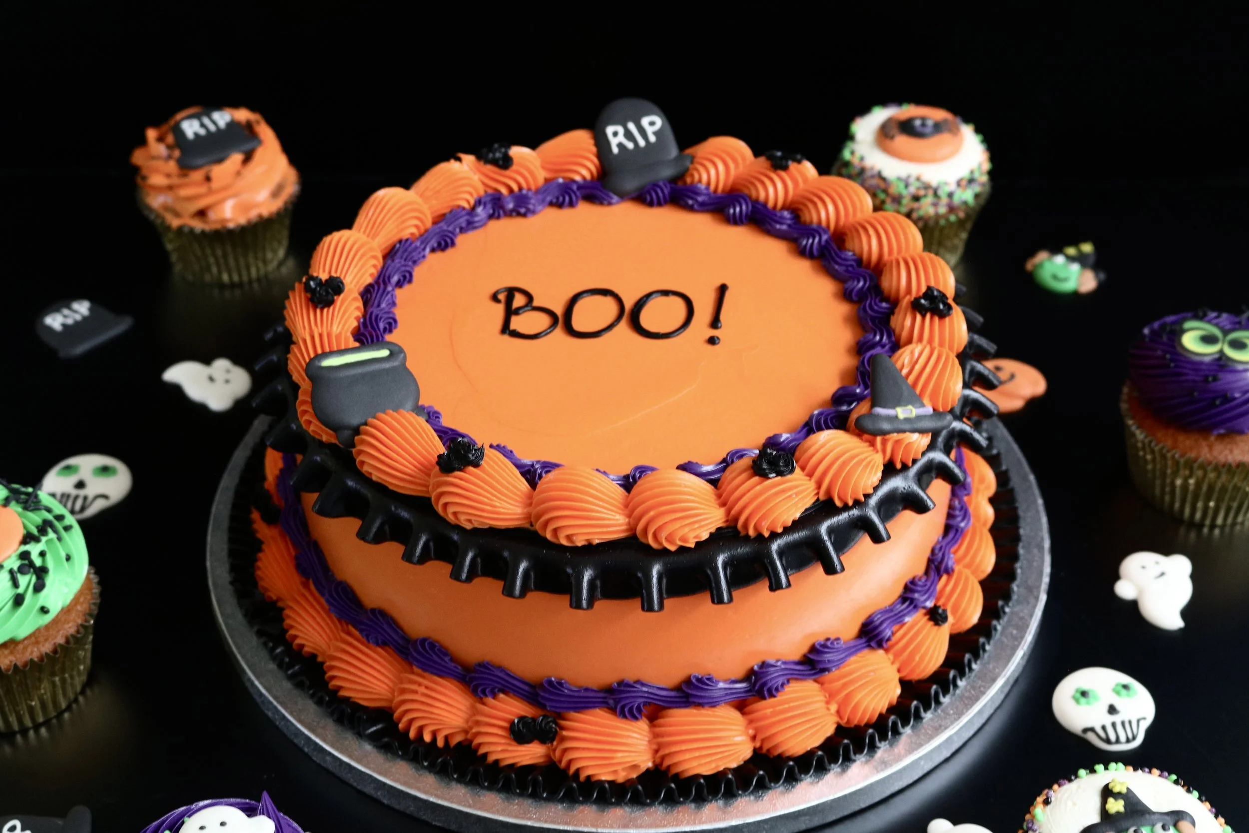Orange vintage Halloween cake with orange and purple piping, black drip, BOO message, cauldron, witch’s hat and RIP gravestone decorations