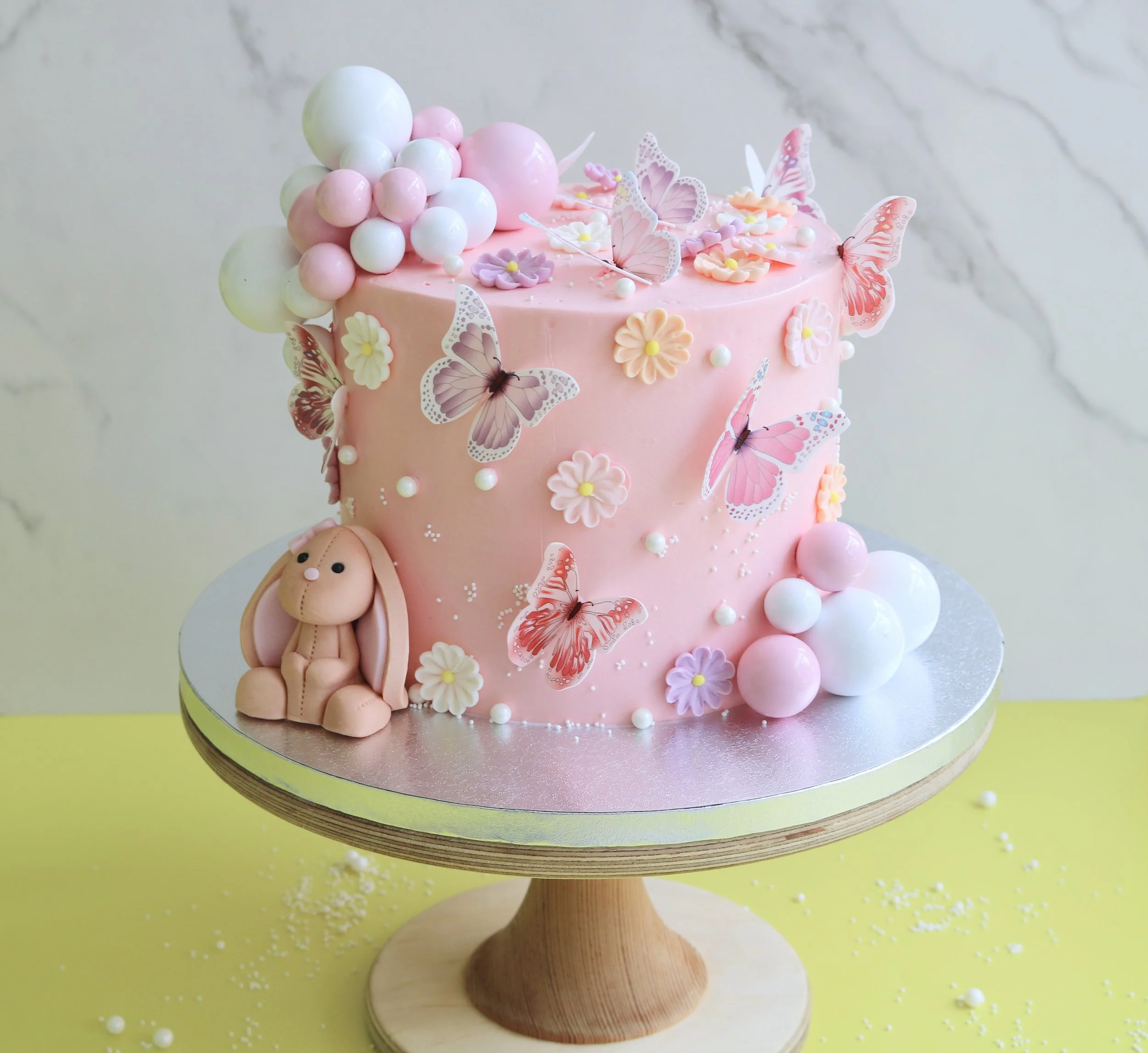 Pastel pink bunny children birthday cake with colourful edible butterflies, edible flowers, pastel balloon decorations, and handcrafted edible bunny topper.
