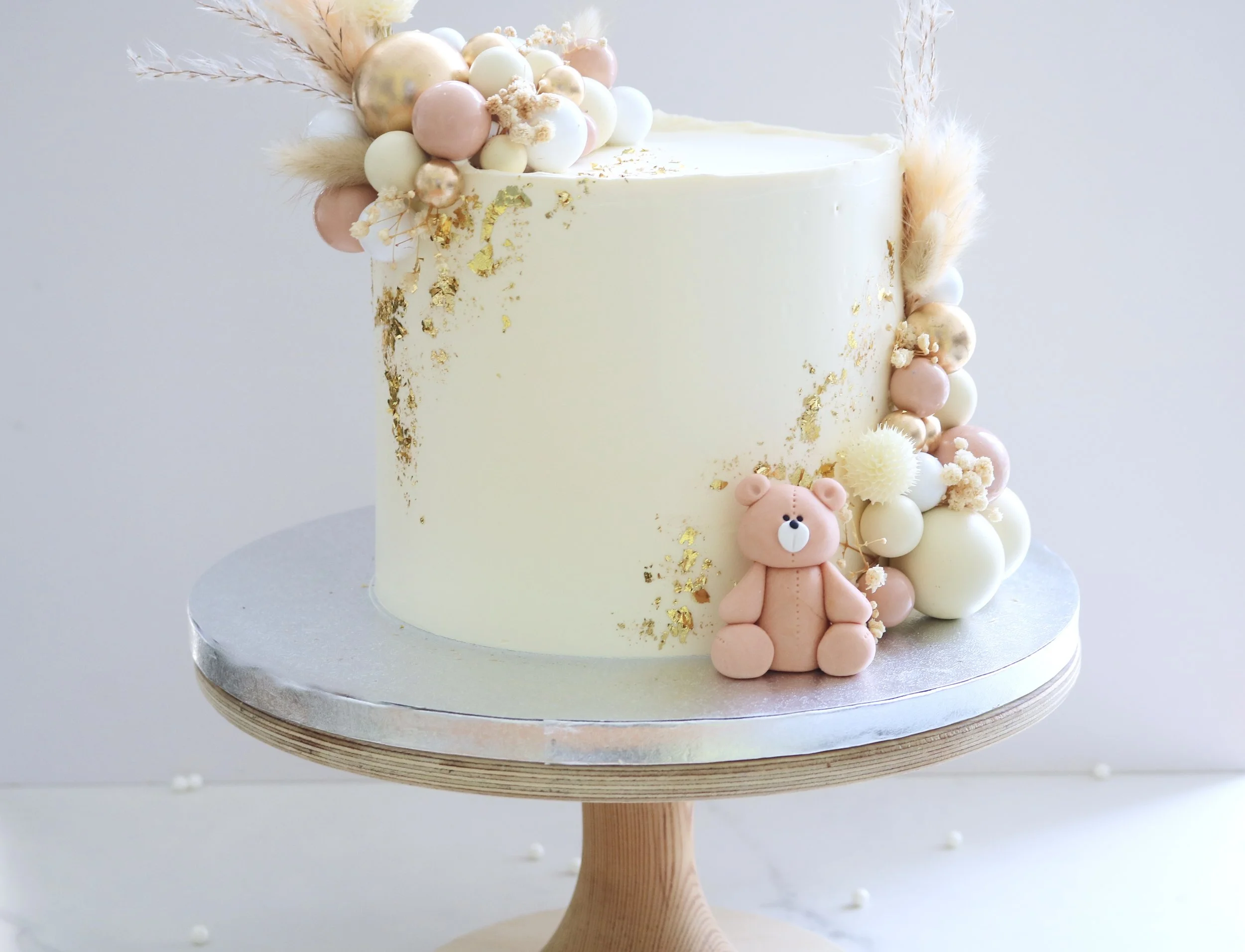 Neutral cream teddy bear children birthday cake with pastel edible balloons, gold leaf decoration, dried floral accents, and handmade edible teddy bear topper.