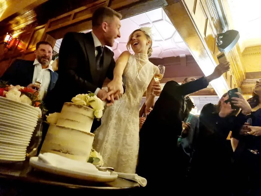 A bride and groom are holding hands and dancing at their wedding reception. The bride is wearing a white dress and holding a glass of wine, while the groom is in a dark suit. Guests are surrounding them, some taking photos, and there is a wedding cake with flowers on the table.