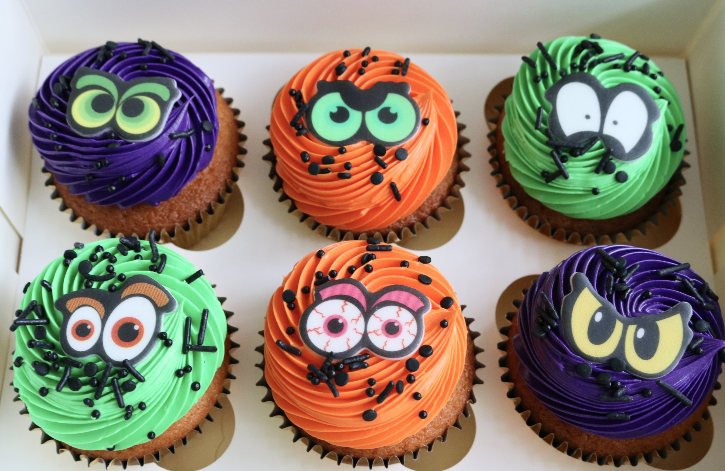 Six Halloween cupcakes with purple, orange and green buttercream swirls, black sprinkles and colourful cartoon monster eye toppers