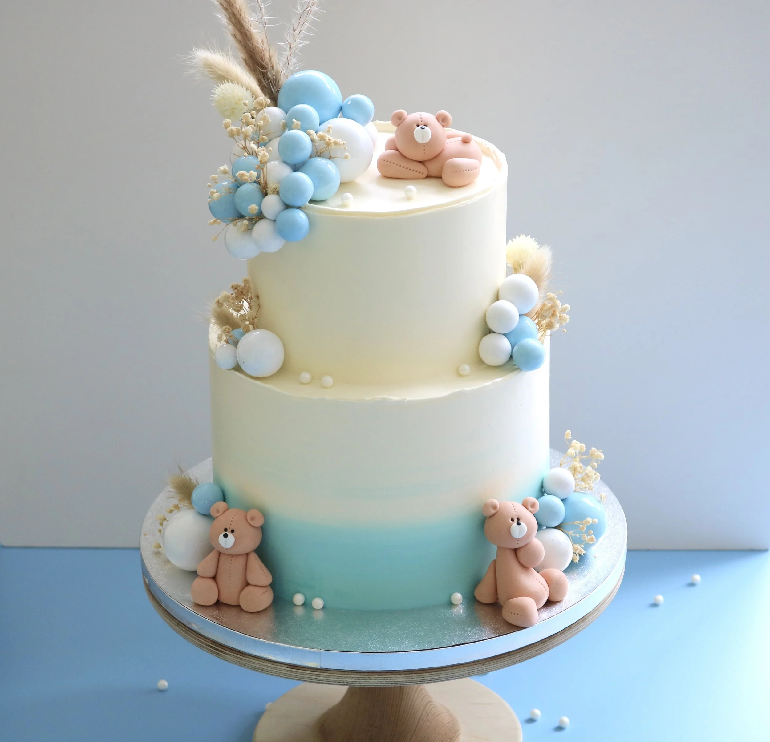 Cream and blue baby cake with handmade edible teddy bears, dried foliage, and balloon decorations, perfect for baby showers, christenings, and first birthdays