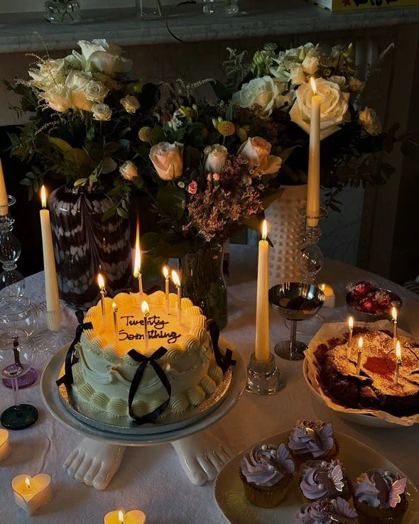 A birthday cake with lit candles on a decorative stand, surrounded by flowers, candles, and desserts on a decorated table.
