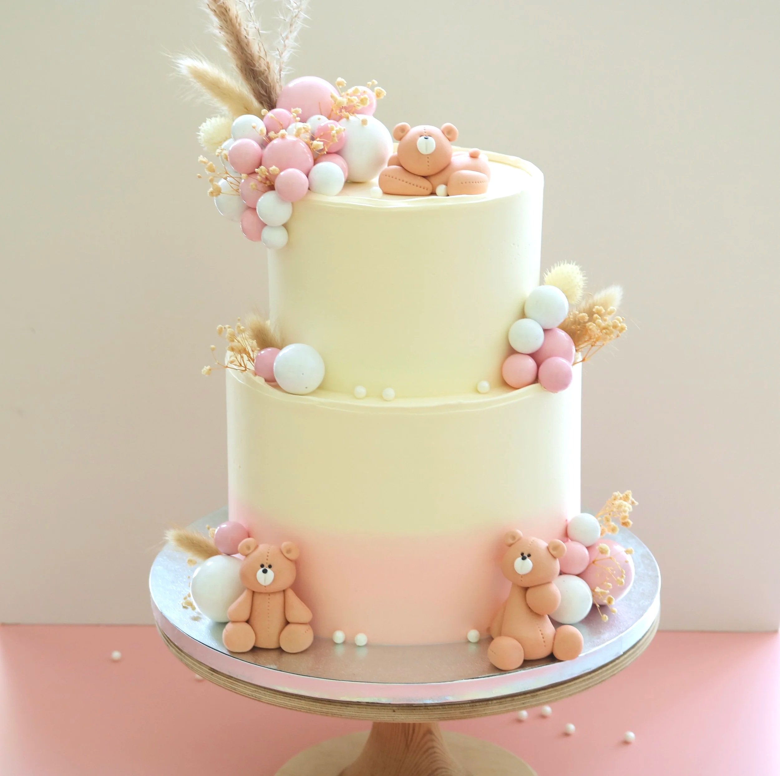 Cream and pastel pink baby cake with handmade edible teddy bears, dried foliage, and balloon decorations, perfect for baby showers, christenings, and first birthdays.