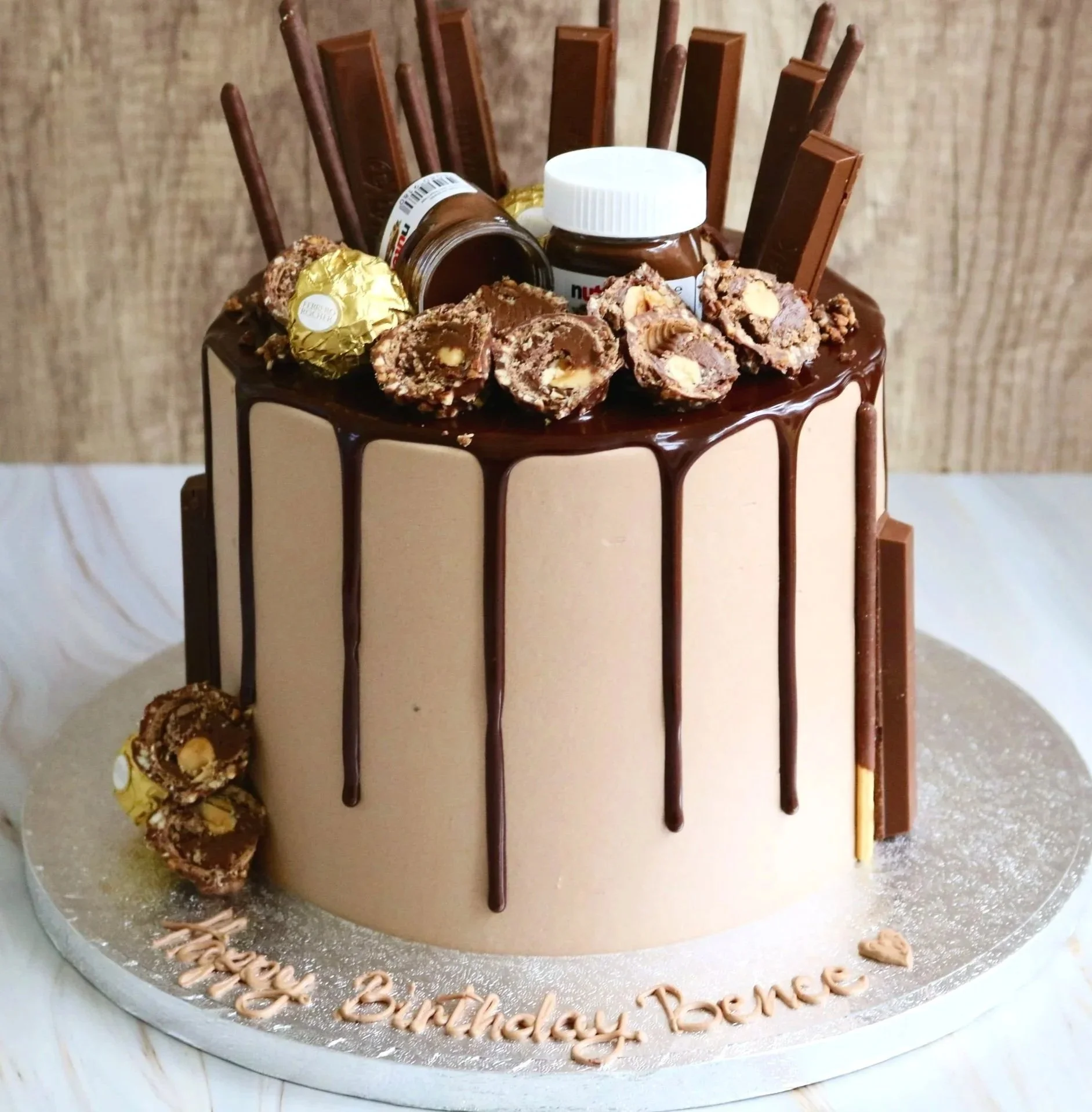 Elegant birthday cake with white, black, and gold marble pattern, decorated with chocolate drip, gold and black macarons, gold-foil accents, and a large chocolate decoration on top. The cake is on a white cake stand with a wooden base, with 'Happy Birthday Nisa' written in chocolate on the base.