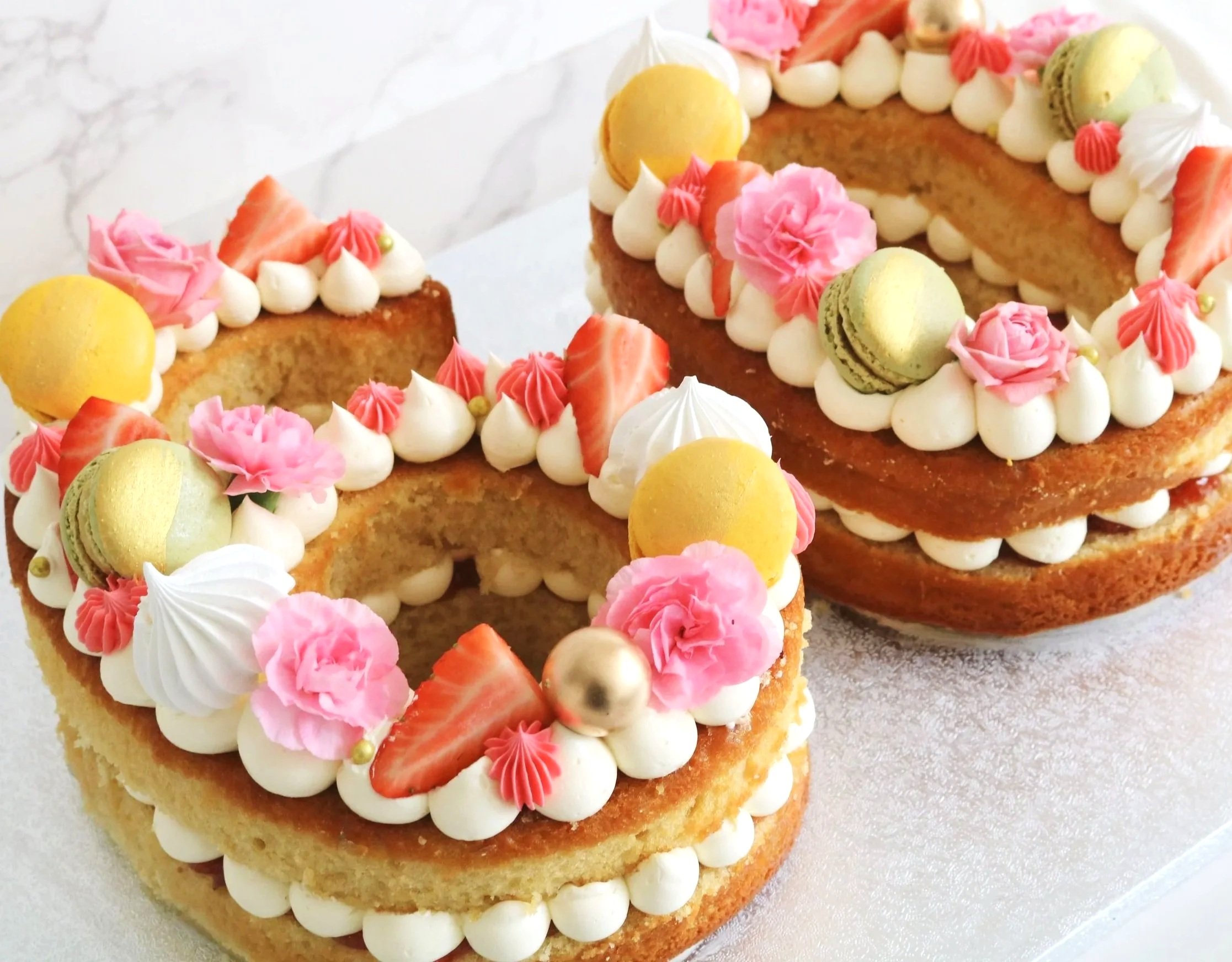Two letter-shaped cakes decorated with pink roses, strawberries, macarons, meringue kisses, and dollops of white frosting, placed on a white surface with a marble background.